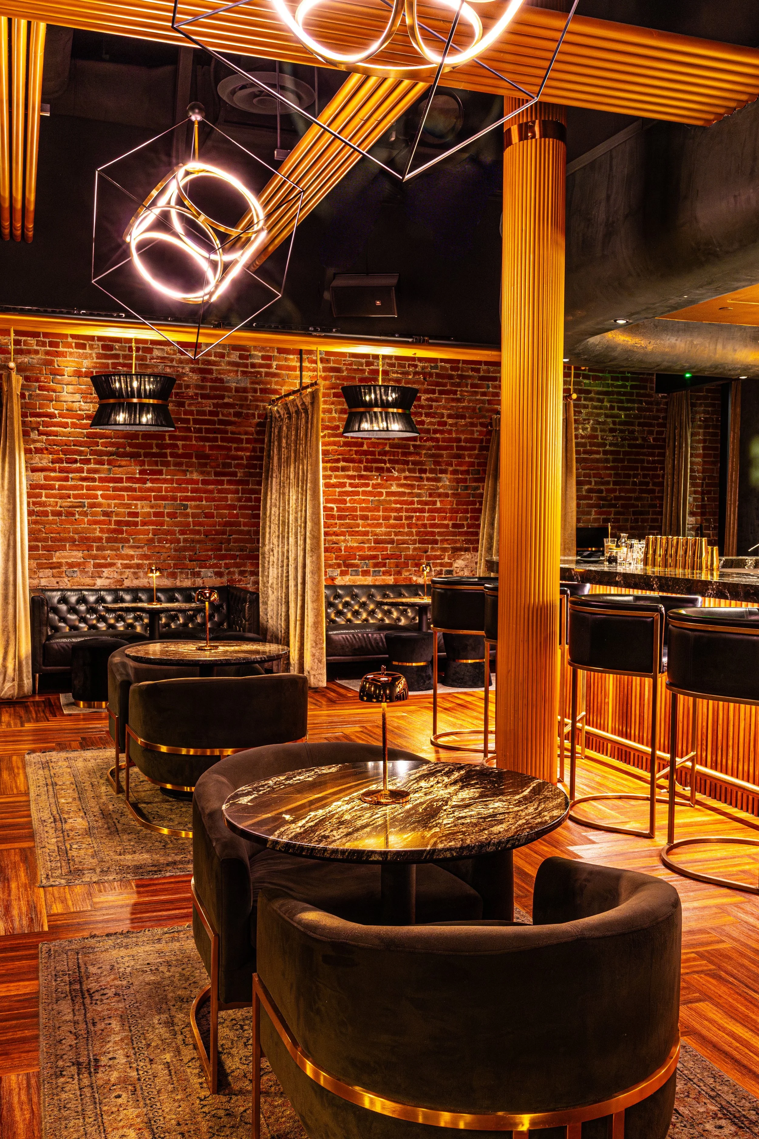 Interior of a stylish bar with black leather chairs, round marble tables, exposed brick wall, gold accents, modern pendant lights, and wood decor elements.