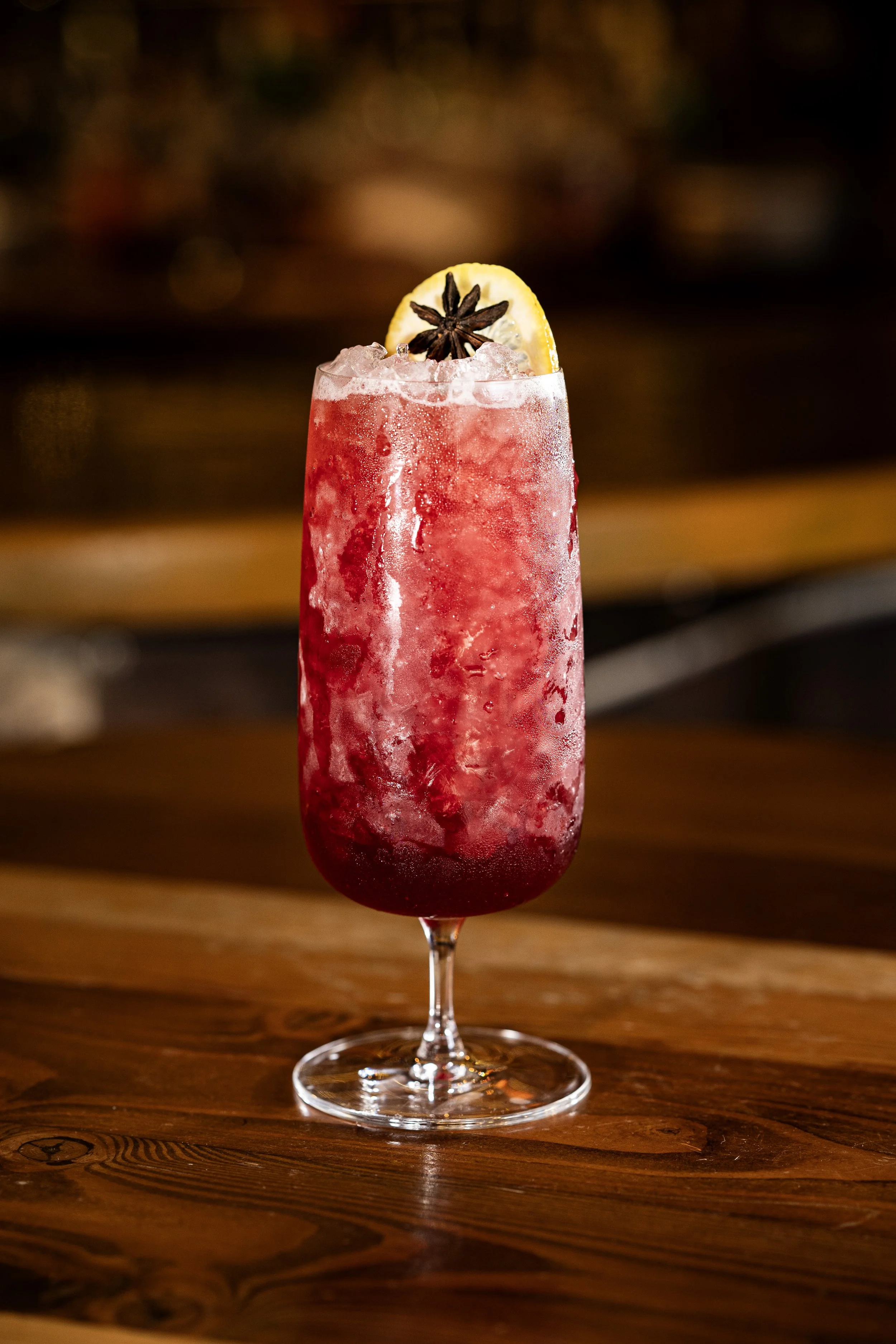 A tall glass filled with a red, icy cocktail garnished with a lemon wedge and a star anise pod, sitting on a wooden surface.