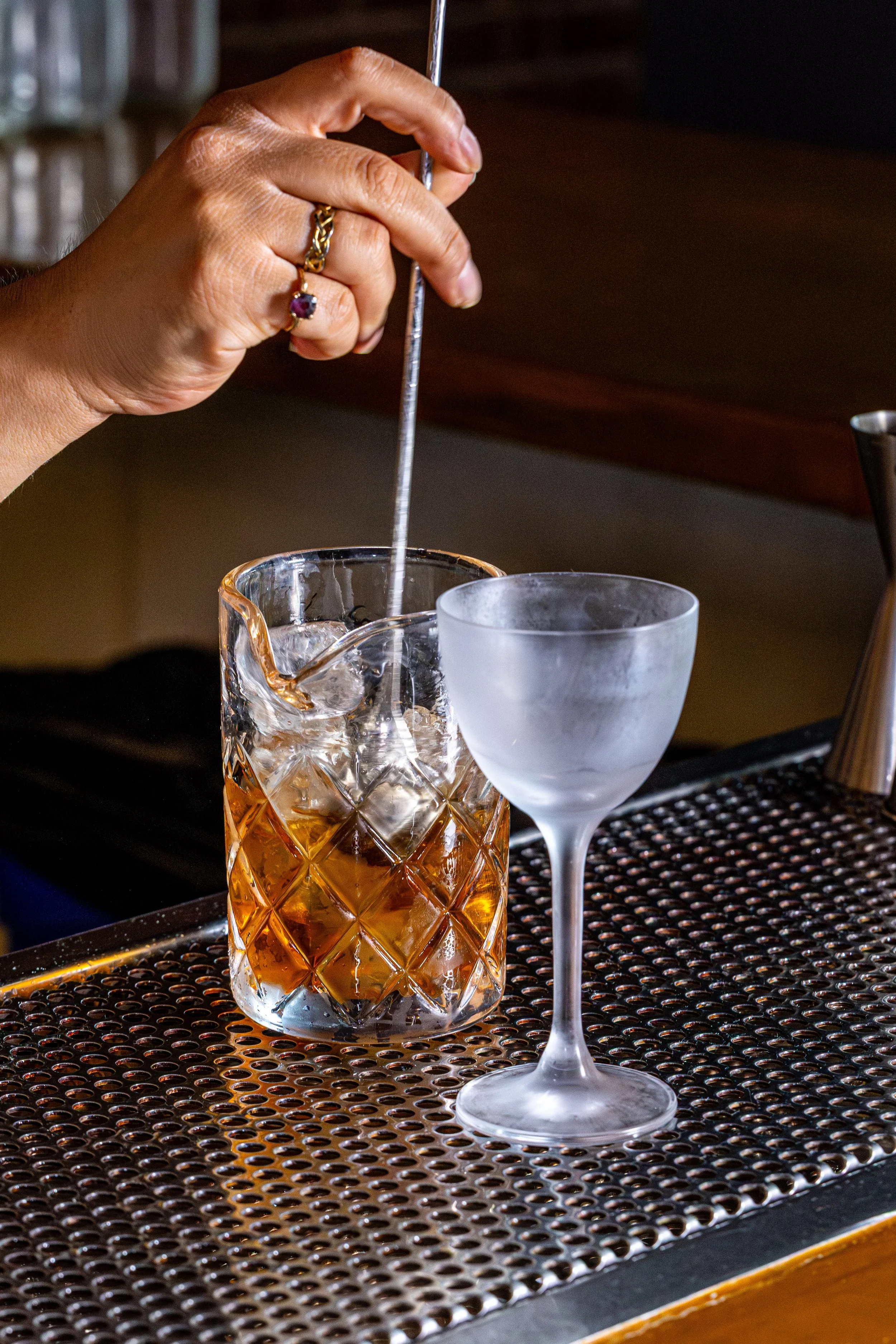 Close-up of a hand stirring a cocktail with a metal bar spoon in a crystal glass on a bar counter. There is also an empty frosted cocktail glass and a bar mat.
