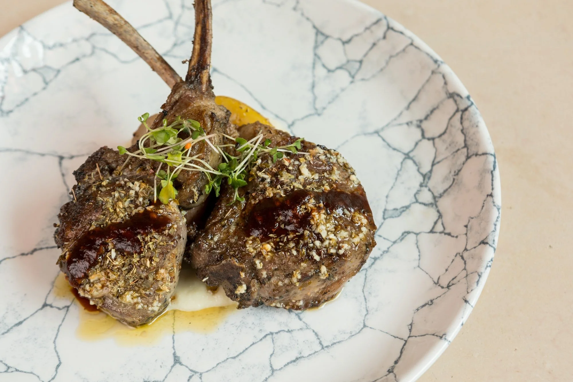 Close-up of four cooked lamb chop pieces topped with herbs, sauce, and microgreens on a white marble plate with dark veining.