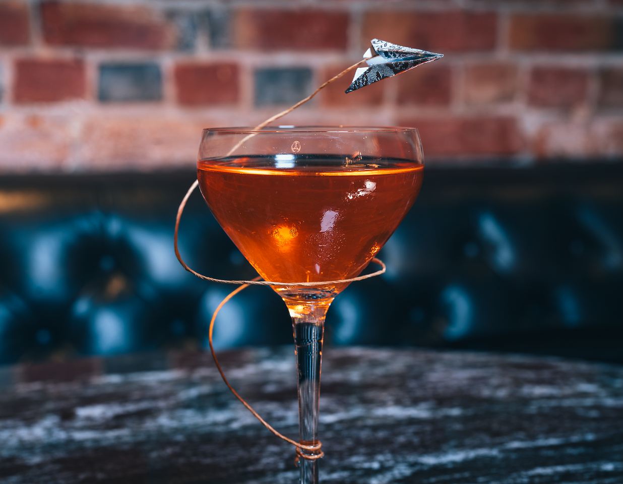 A coupe glass filled with a golden amber drink, topped with a small paper airplane on a stick as a decorative garnish, placed on a dark wooden surface with a brick wall in the background.