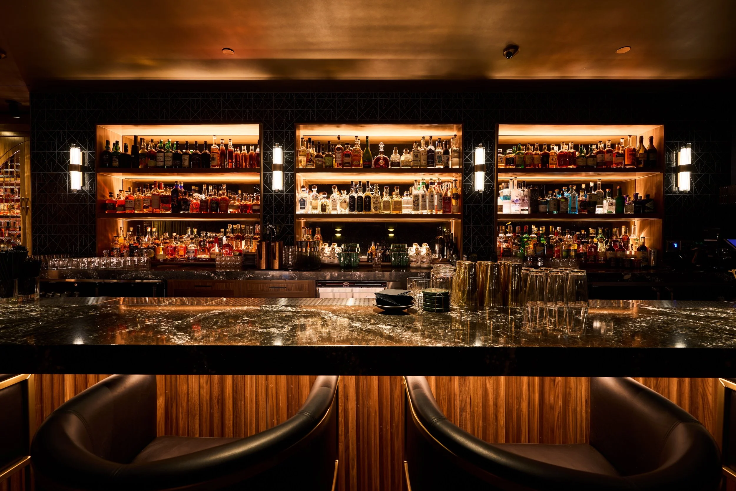 A bar with illuminated wooden shelves filled with various bottles of alcohol, two vertical light fixtures on each side, a marble countertop, and black bar stools in the foreground.