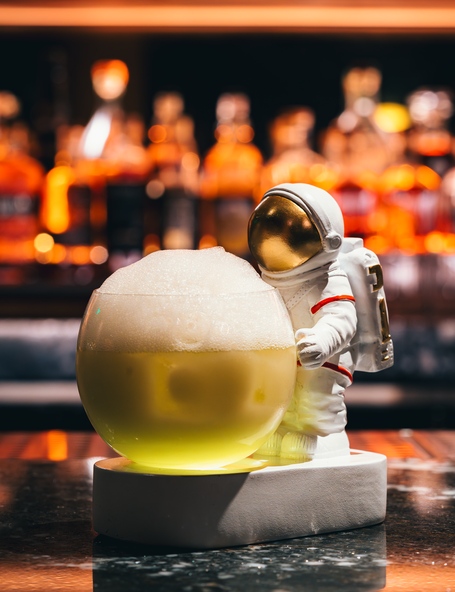 A toy astronaut figure standing beside a large round yellow object with foam on top, set against a blurry background of orange and black lights.