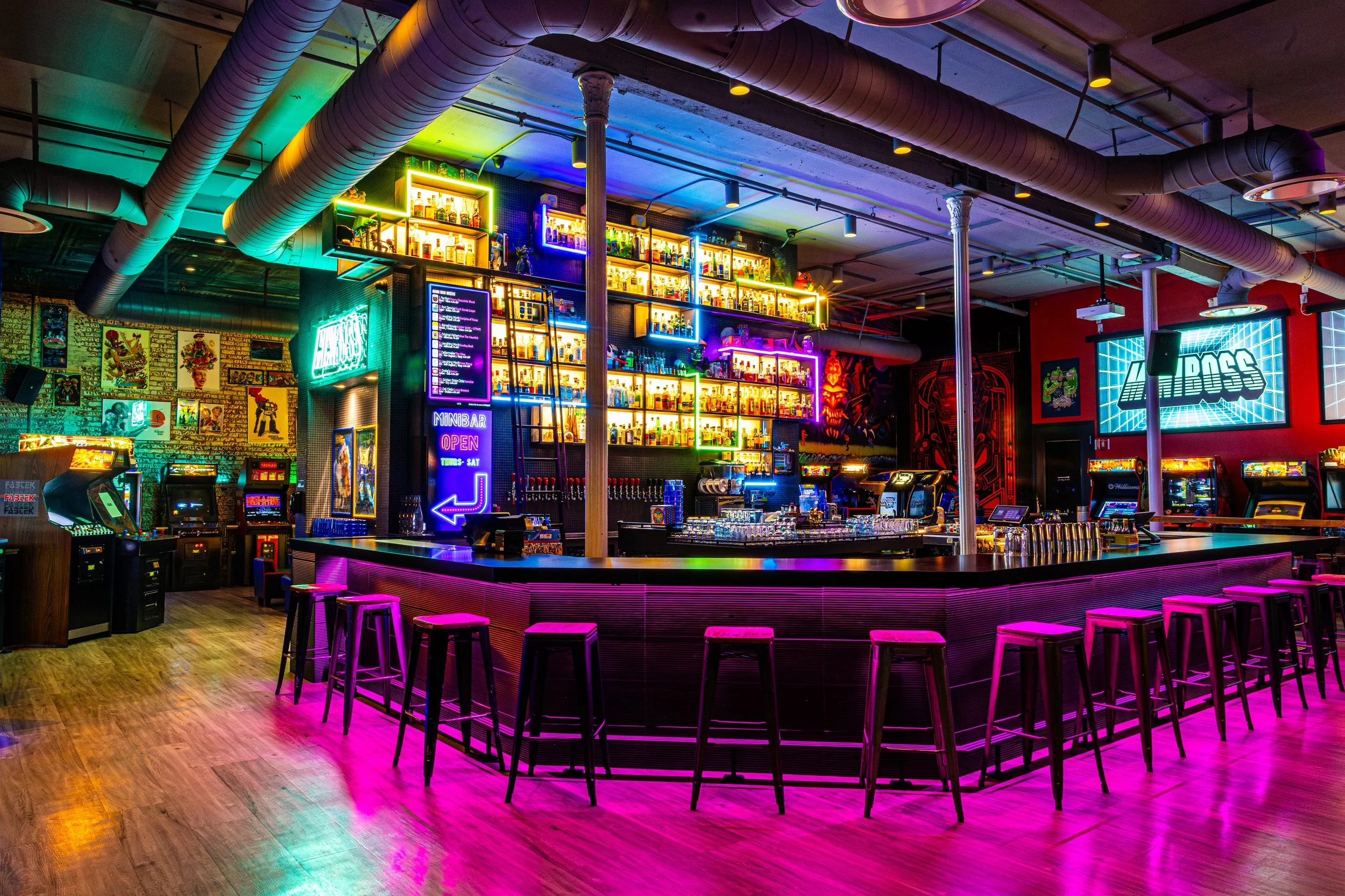 A vibrant bar area with colorful LED lighting, shelves stocked with various bottles of alcohol, and arcade machines. The space has a modern, neon-lit decor with purple, yellow, and green accents, and a wooden floor.