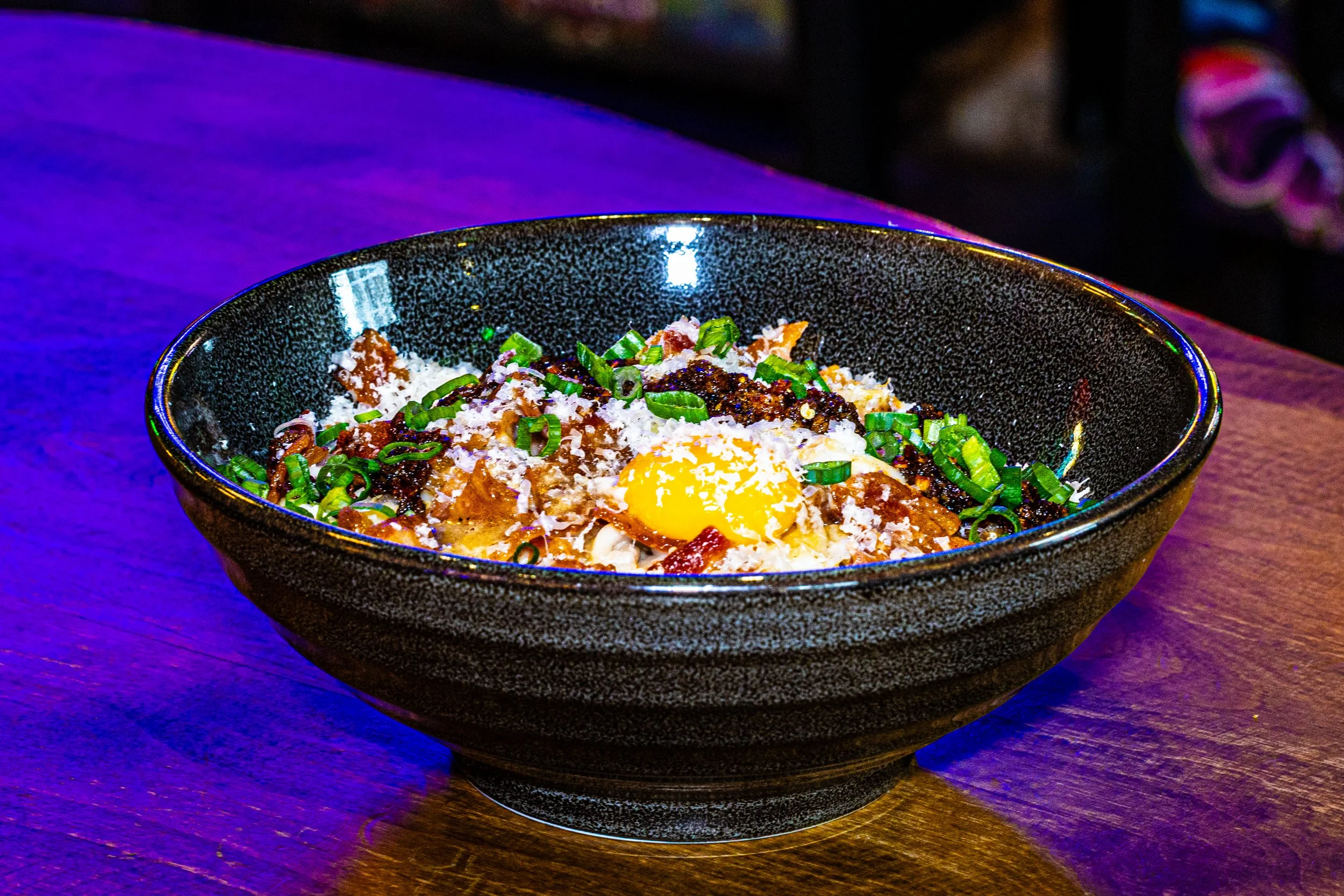 A black bowl containing rice, ground meat, a cooked egg with a runny yolk, chopped green onions, and grated cheese, placed on a wooden surface with purple lighting.