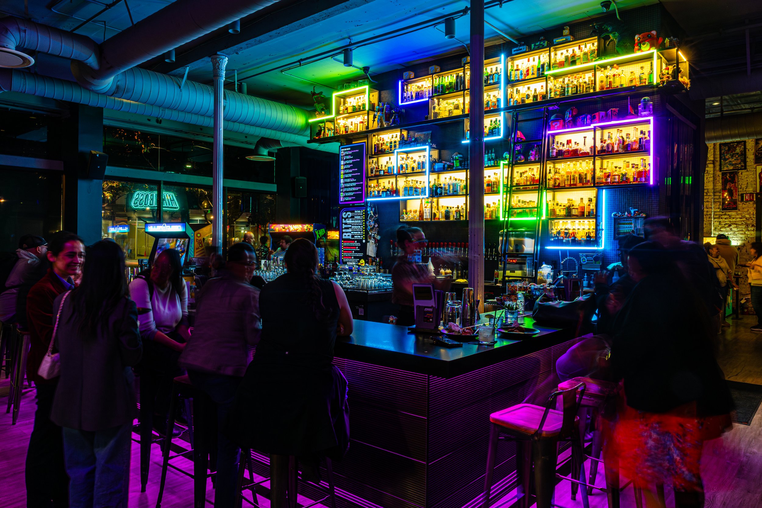 A busy bar with shelves of liquor bottles illuminated by colorful neon lights. People sit and stand around the bar, with some enjoying drinks and socializing in a lively atmosphere.
