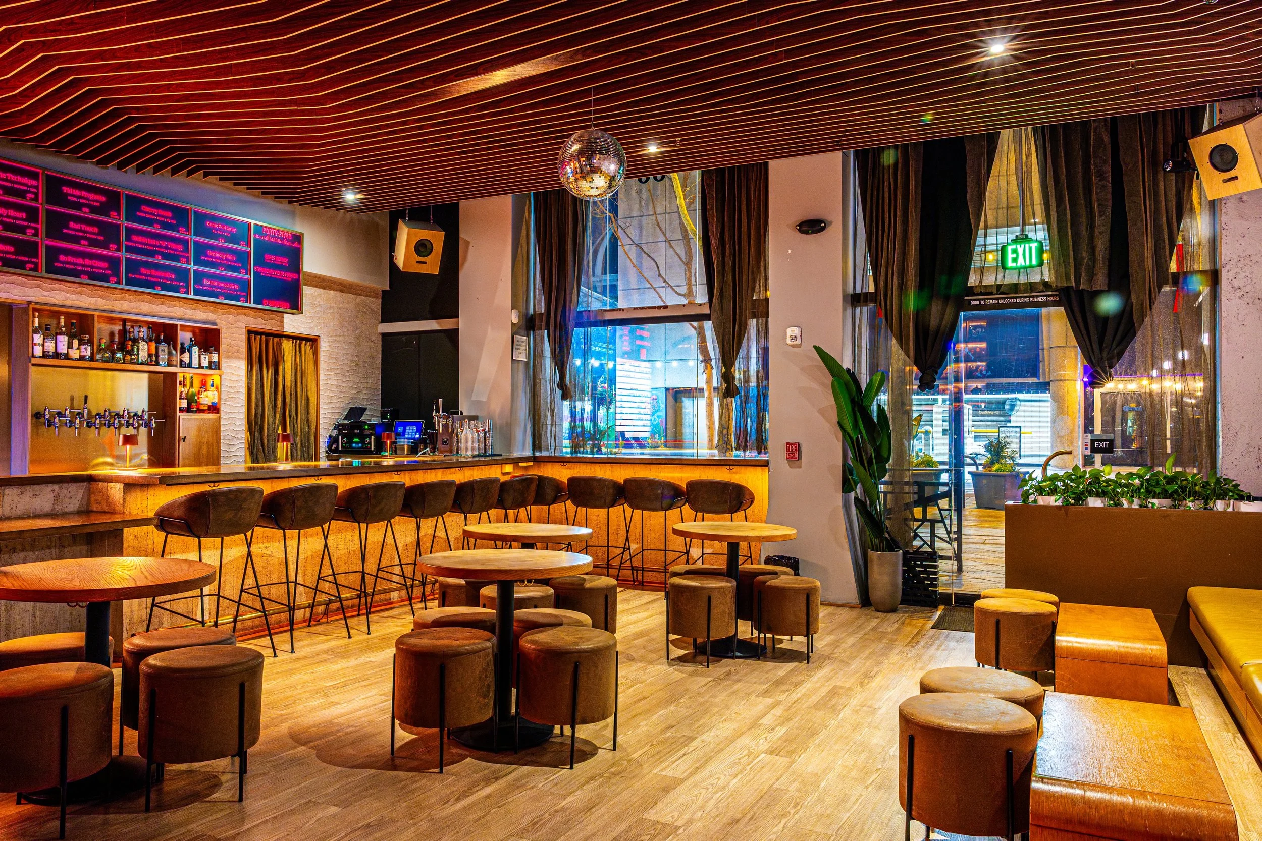 Interior of a modern bar and lounge area with a wooden bar counter, bar stools, tables, stools, plants, large windows with curtains, a disco ball hanging from the ceiling, neon light outdoors, and an exit sign.