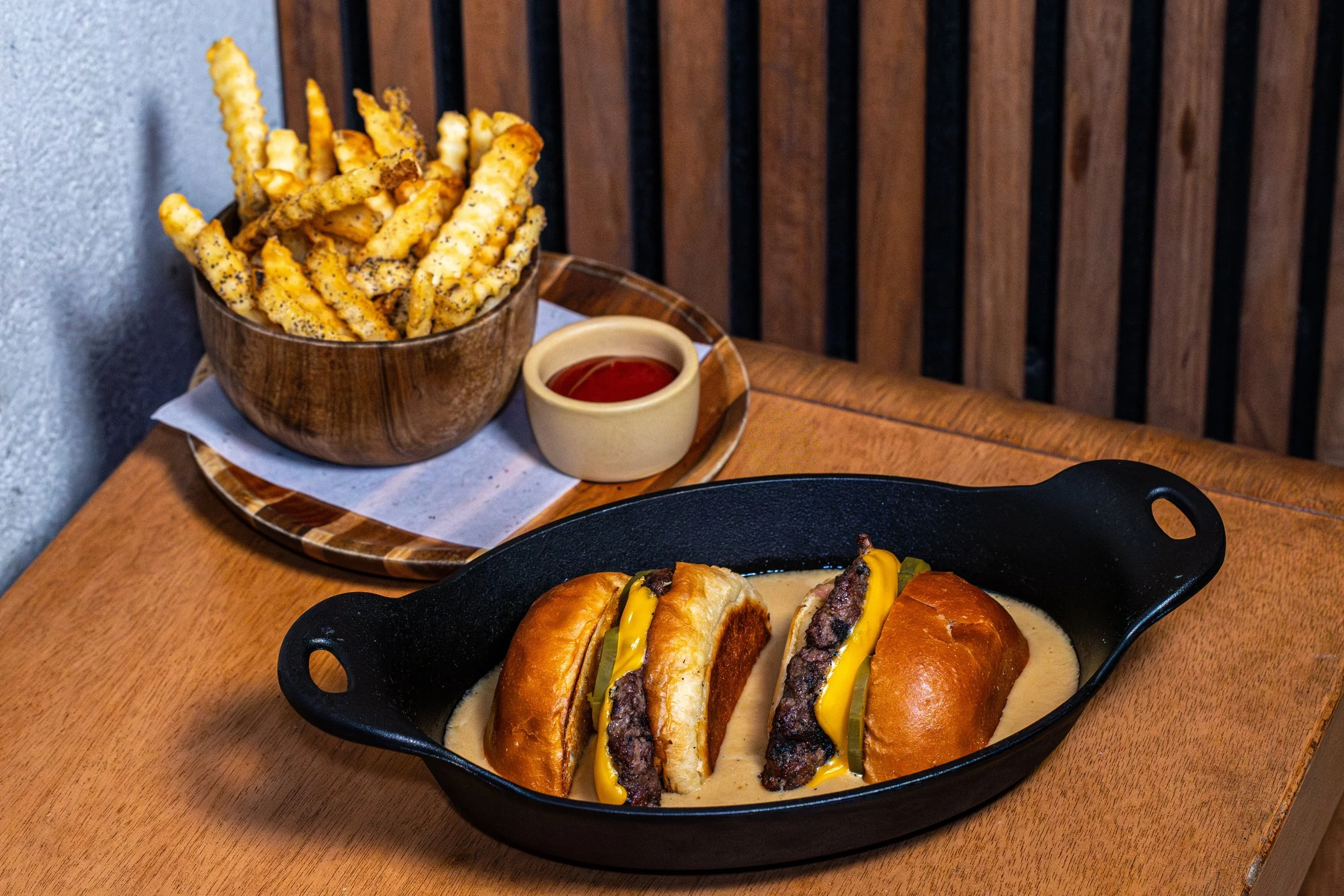 A bowl of crinkle-cut French fries with black pepper seasoning, served with ketchup in a small cup, on a wooden tray. In the foreground, a black skillet with hot dogs topped with sliced pickles and mustard.