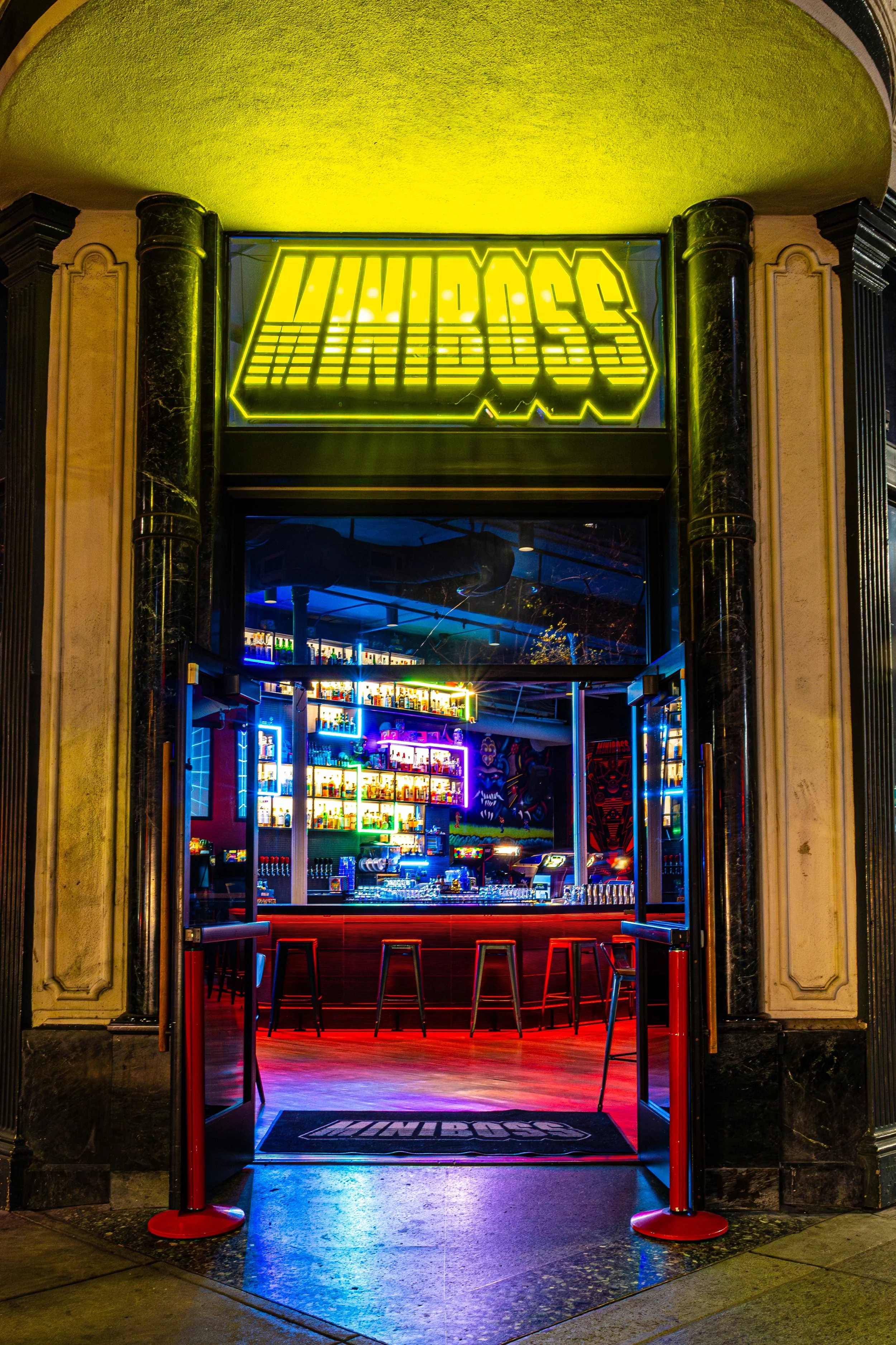 Entrance to a bar with a neon sign that reads 'WURDOSS' above the door, illuminated shelves with bottles visible inside, black marble pillars, bar stools, and colorful neon lighting creating a vibrant atmosphere.