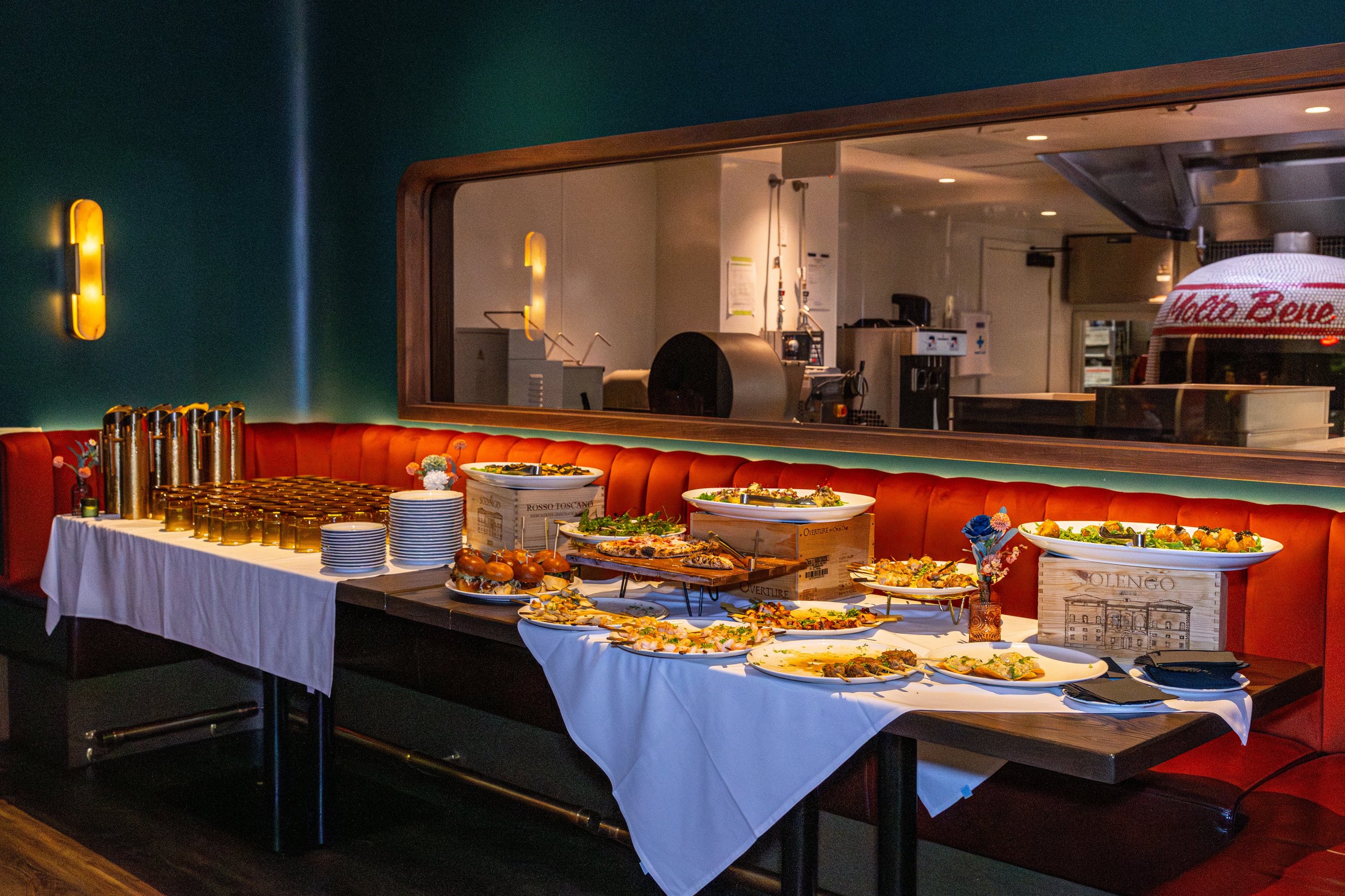 Buffet table with various dishes including salad, pizzas, and sliders, set in a restaurant with a serving window and wall lighting.
