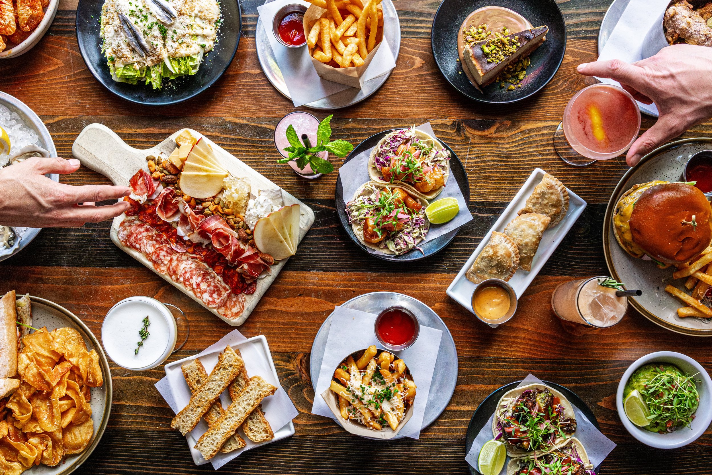 A variety of dishes on a wooden table, including tacos, French fries, cheese and charcuterie platter, salad, and drinks, at a social gathering.