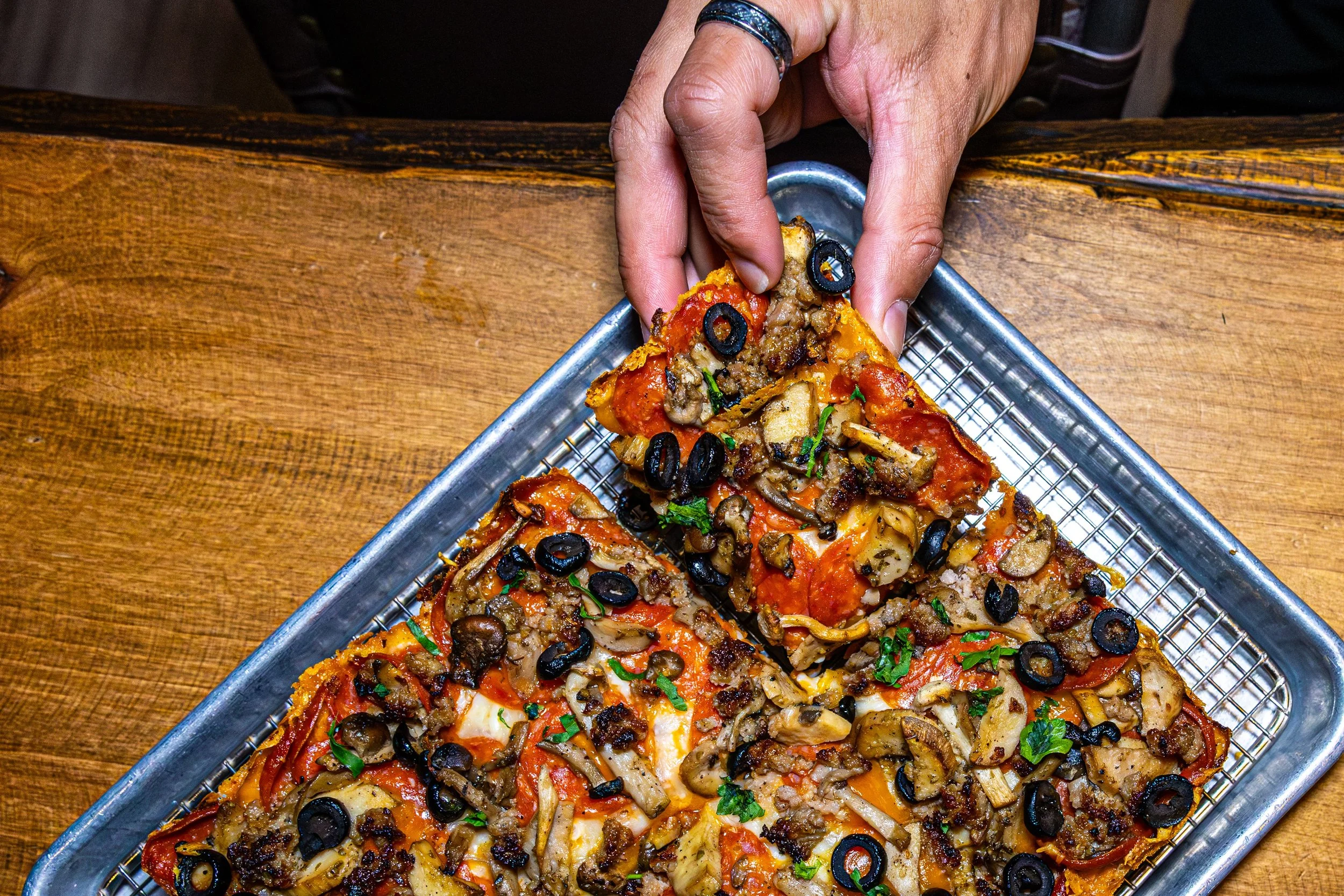 Person placing black olives on sliced pizza with toppings of pepperoni, sausage, mushrooms, and parsley, on a metal tray over a wooden table.
