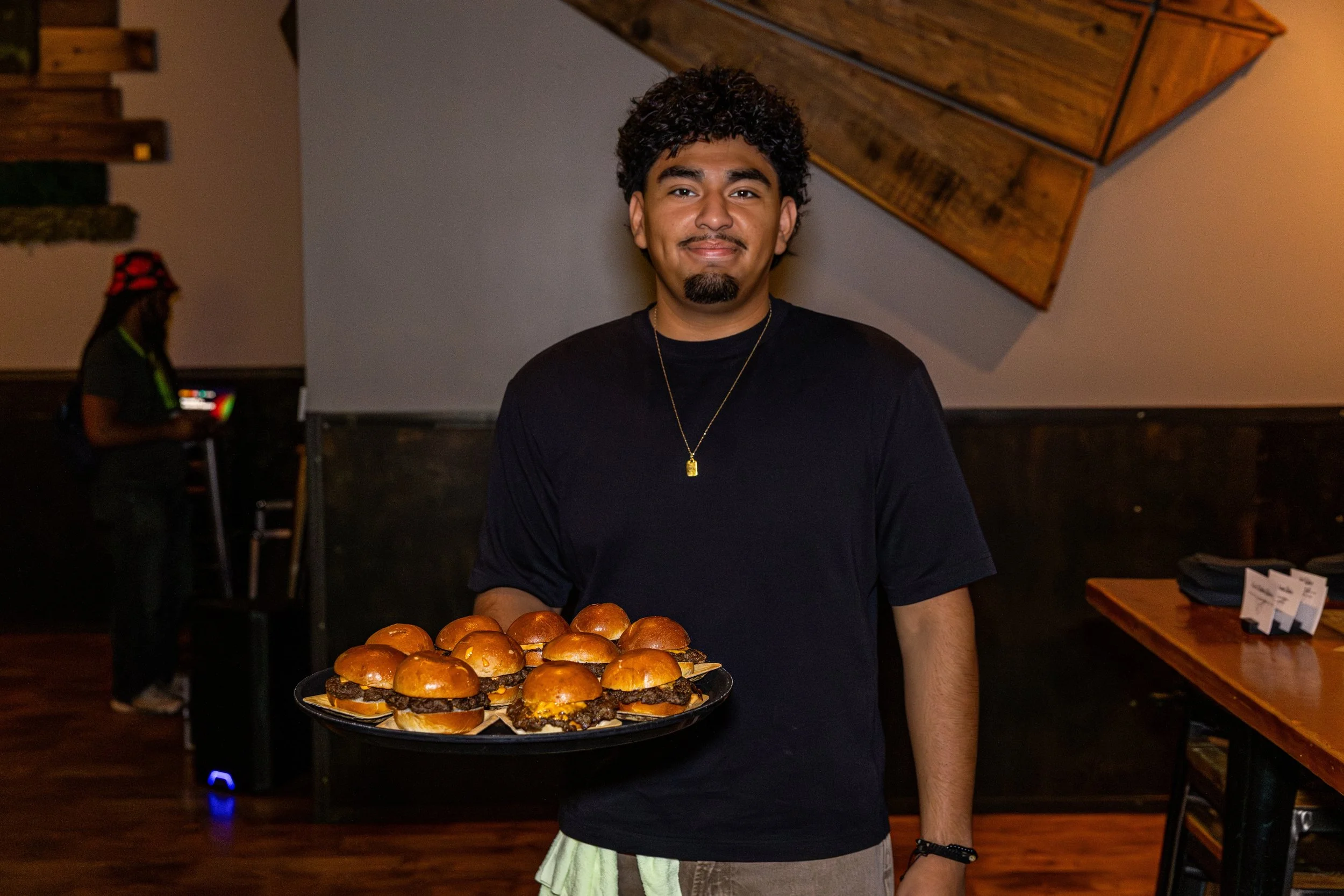 A young man with dark curly hair and a goatee presenting a large tray of sliders with beef patties and buns, standing in an indoor setting with wooden decor.