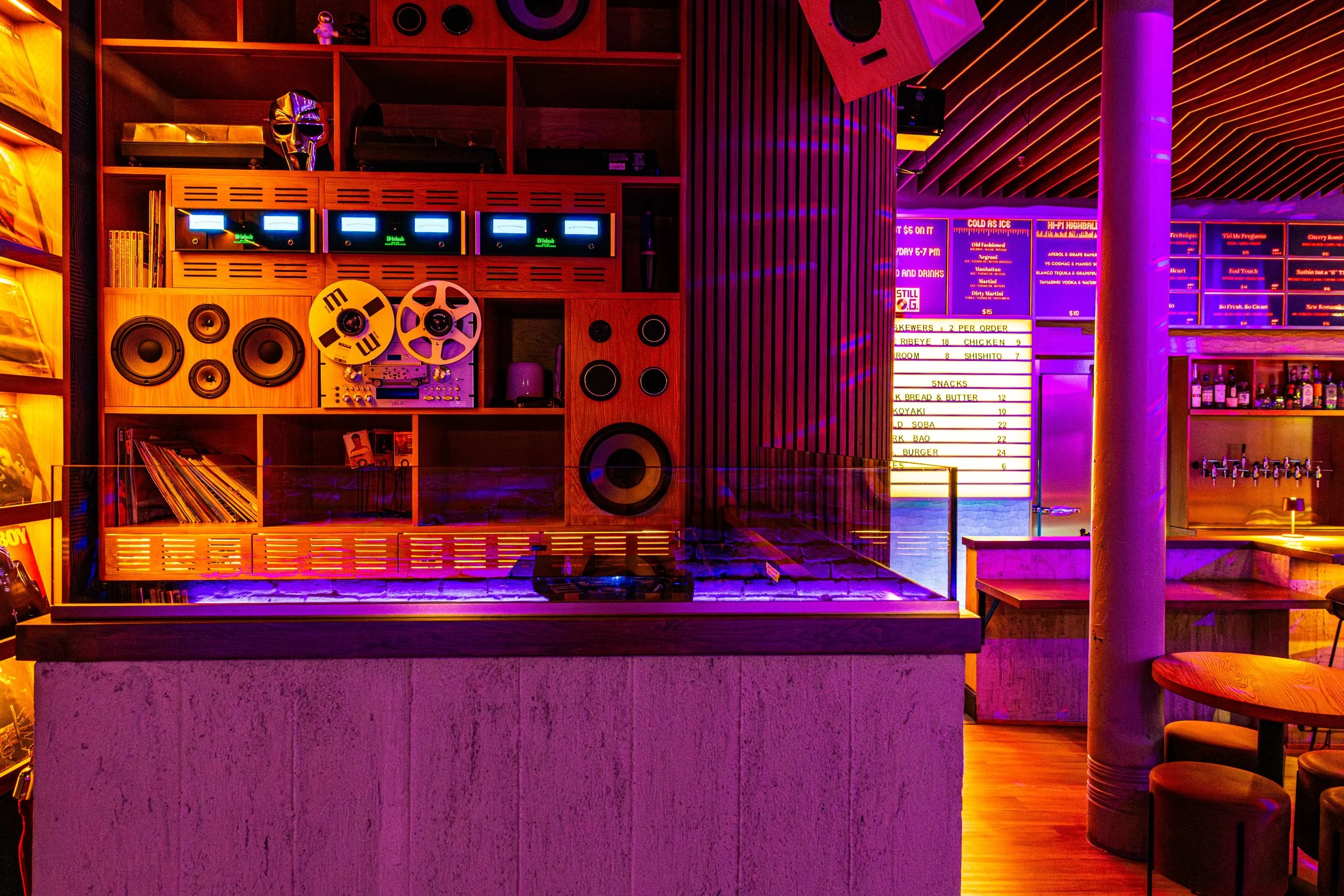 A vibrant interior scene at a bar or nightclub with purple and orange lighting, featuring a wooden wall with vintage audio equipment, vinyl records, a DJ turntable, speakers, and a neon menu sign listing food and drink options.