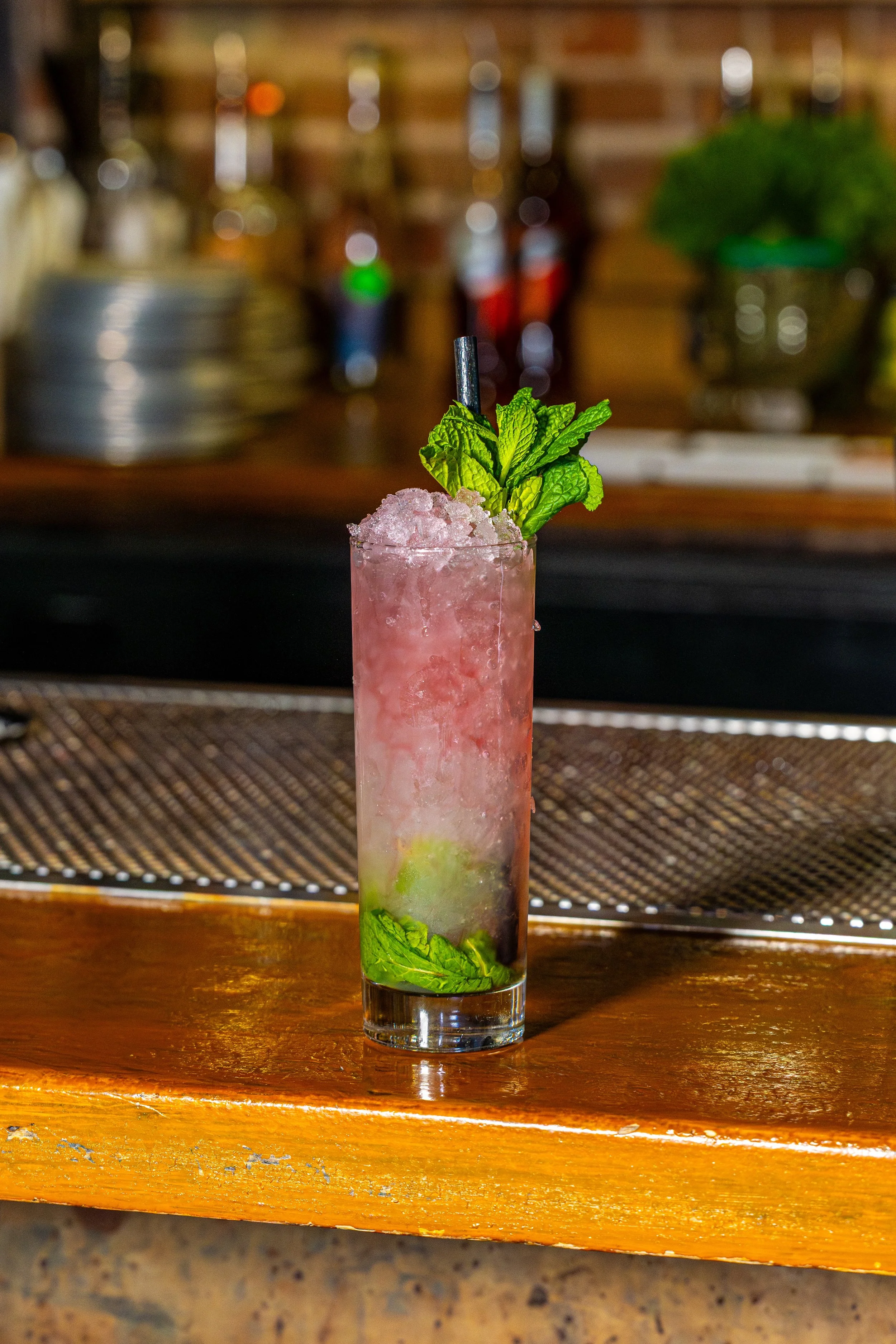 A tall glass filled with a pink, crushed ice beverage garnished with fresh green mint leaves and a black straw. The drink is on a wooden bar counter with a blurred background of bottles and bar equipment.