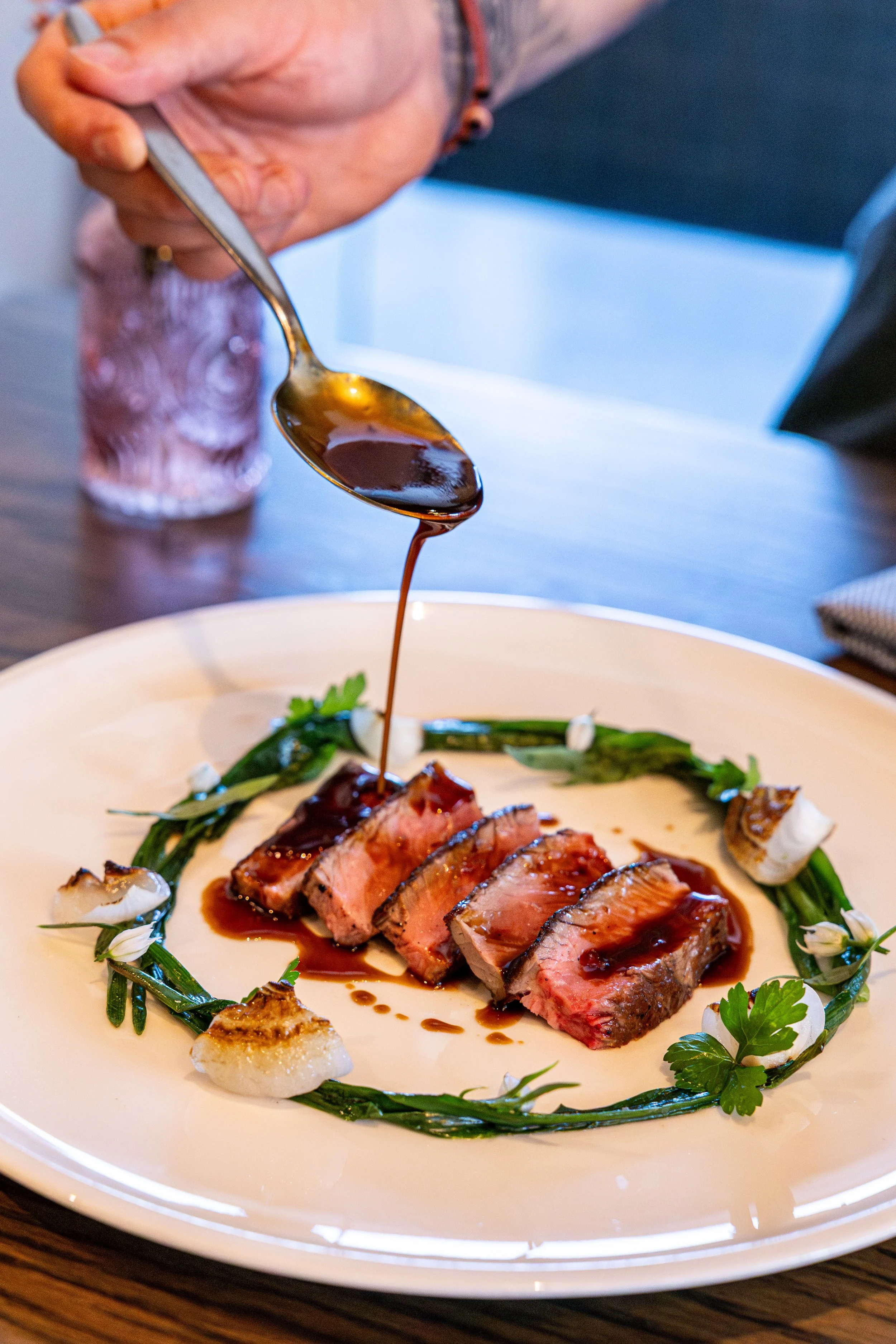 Sliced medium-rare steak with sauce being poured on top, garnished with herbs and vegetables on a white plate.