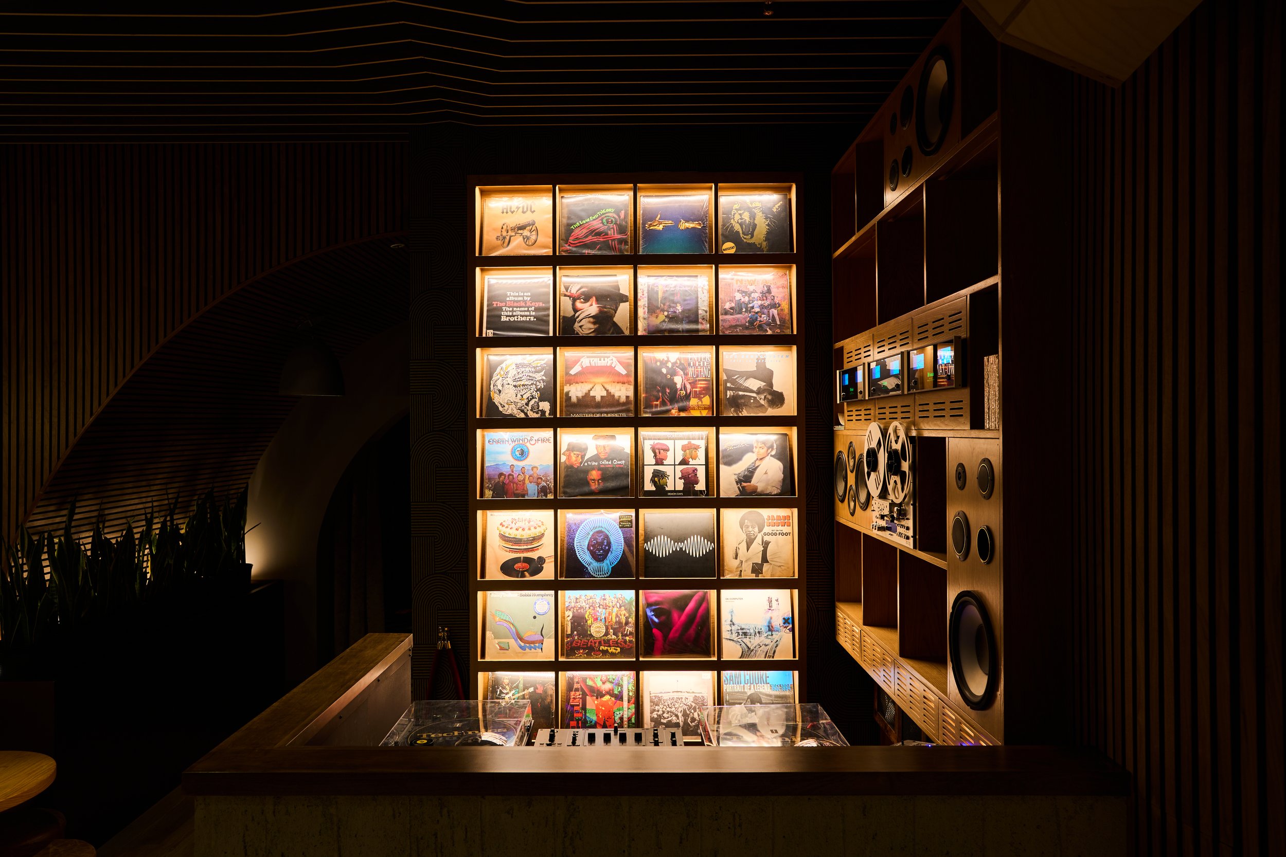 A wall display featuring illuminated album covers and artwork, with a DJ turntable setup in the foreground and speakers on the right, in a dimly lit room with wooden accents.