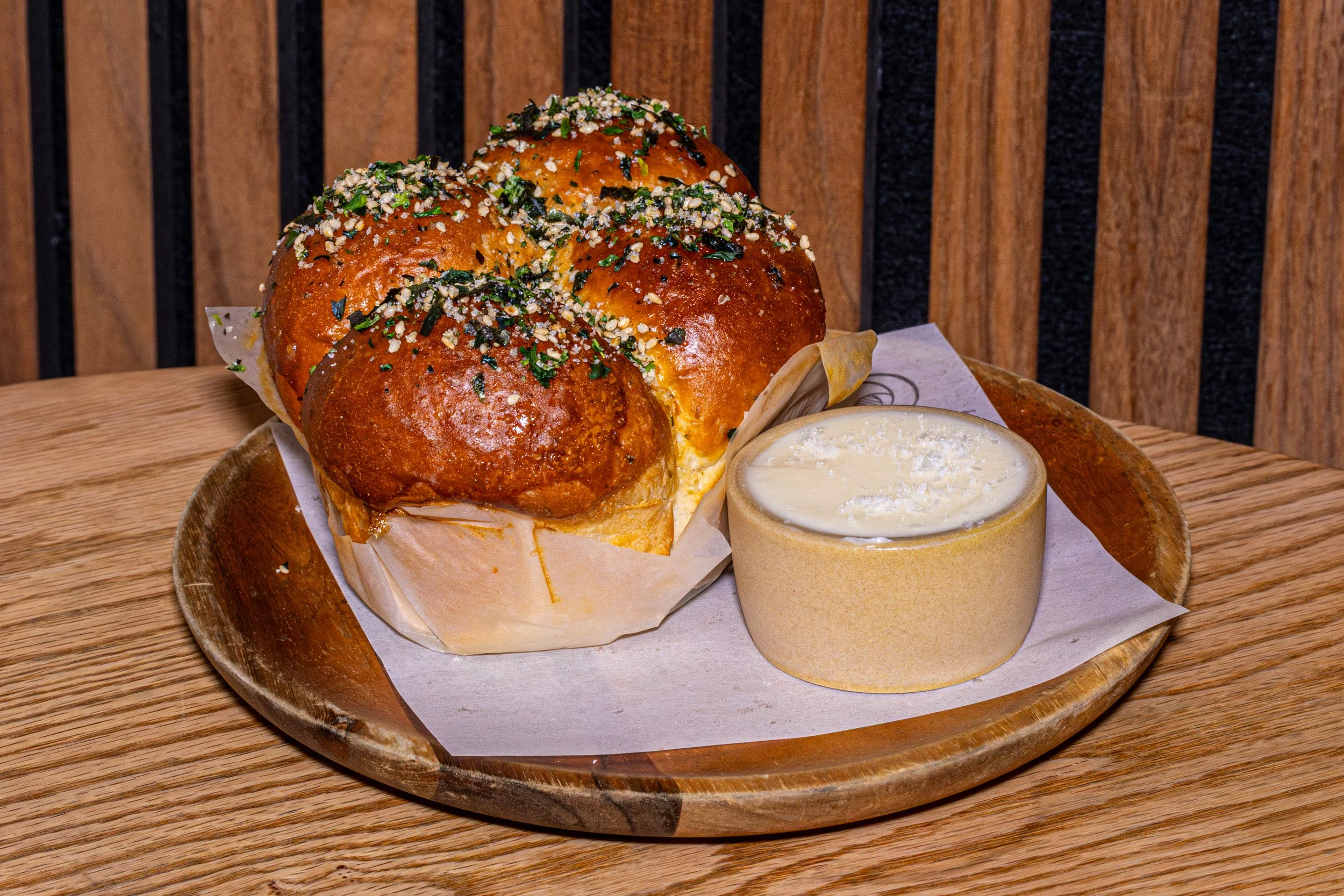 Three brown dinner rolls topped with herbs and sesame seeds, served with a small dish of butter on a wooden plate.