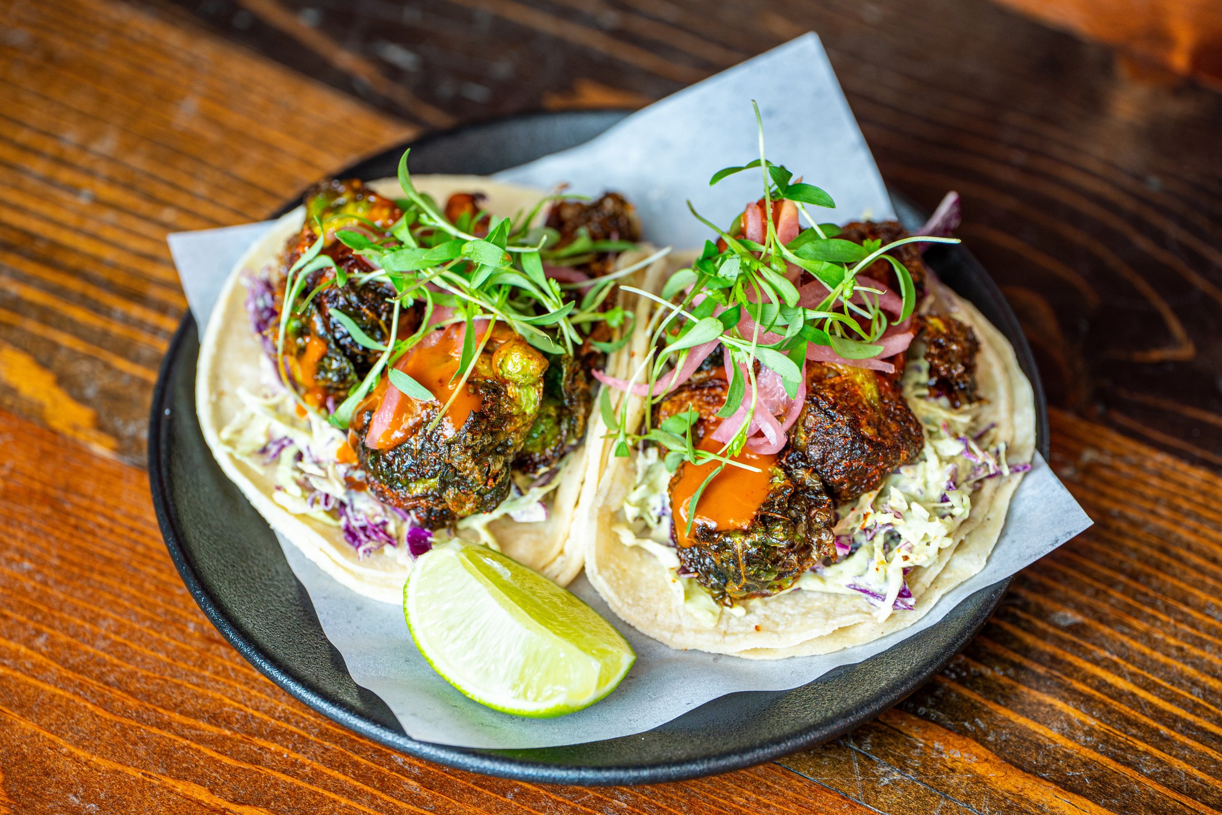 Close-up of two beef tacos filled with seasoned meat, topped with fresh microgreens, and accompanied by lime wedges on a black plate on a wooden table.
