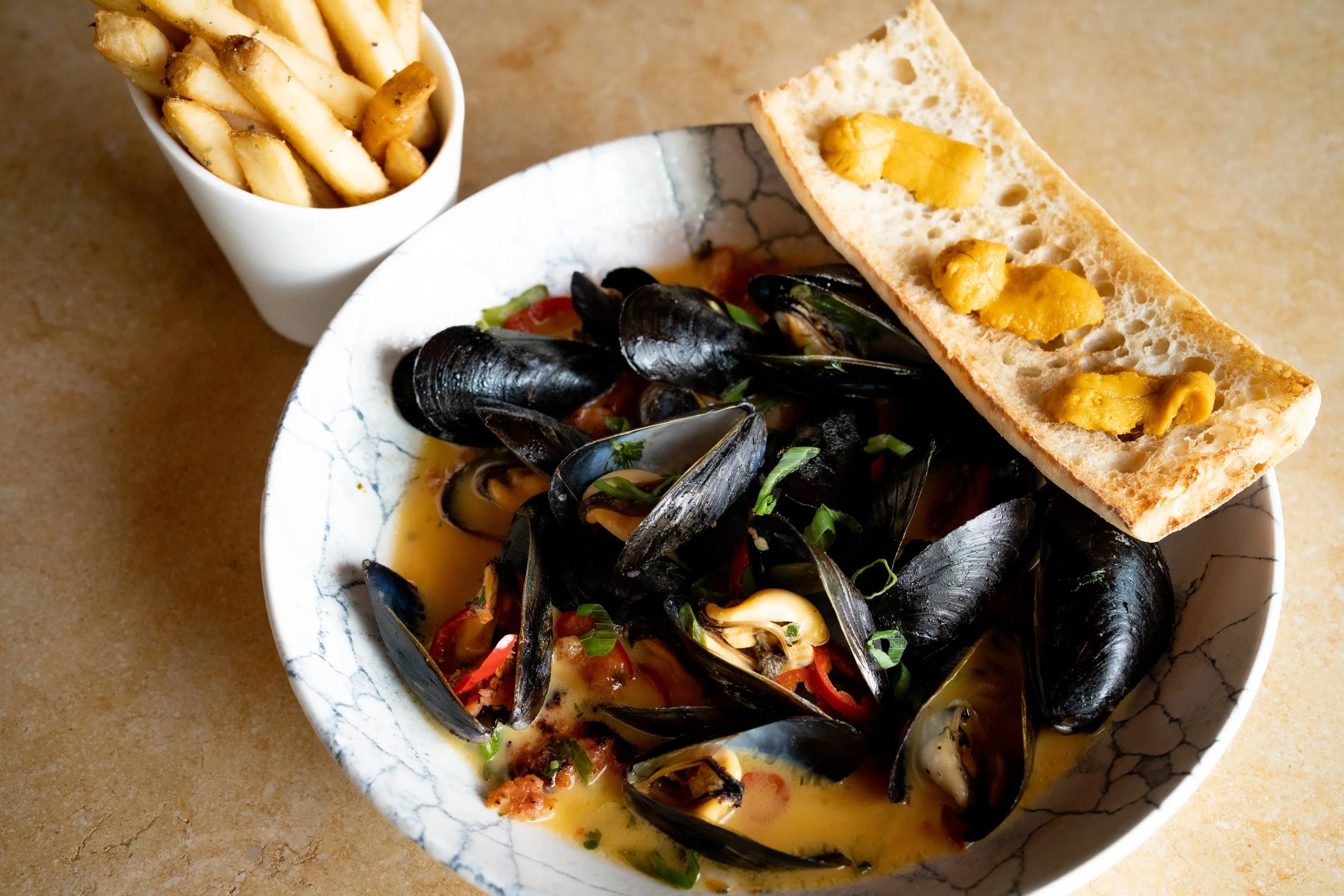 Bowl of cooked mussels with chopped herbs and sliced red peppers, with slices of toasted bread topped with yellow mustard on the side.