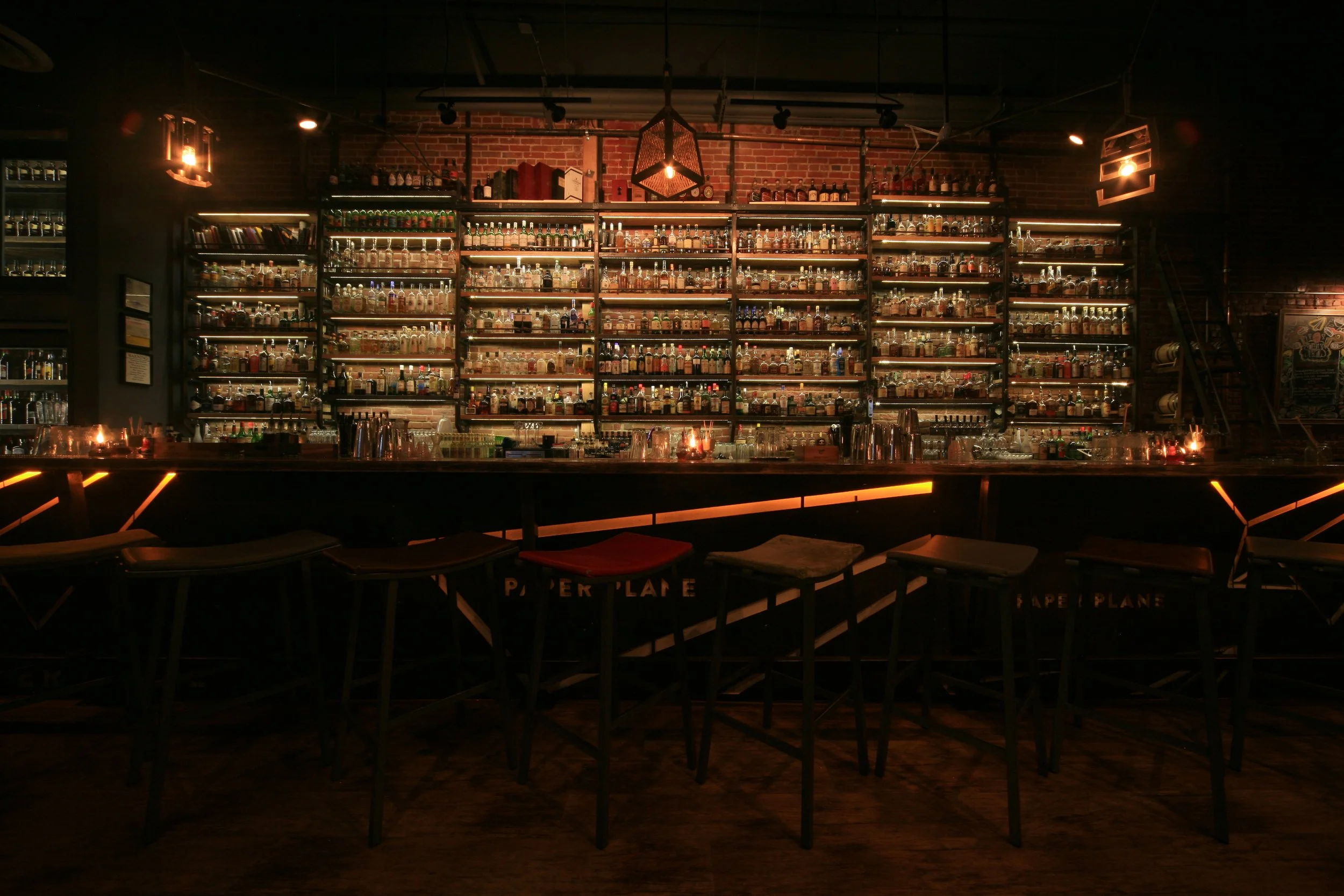 A bar with a large collection of liquor bottles displayed on shelves against a brick wall, dim lighting with hanging fixtures, and a bar counter with stools in the foreground.