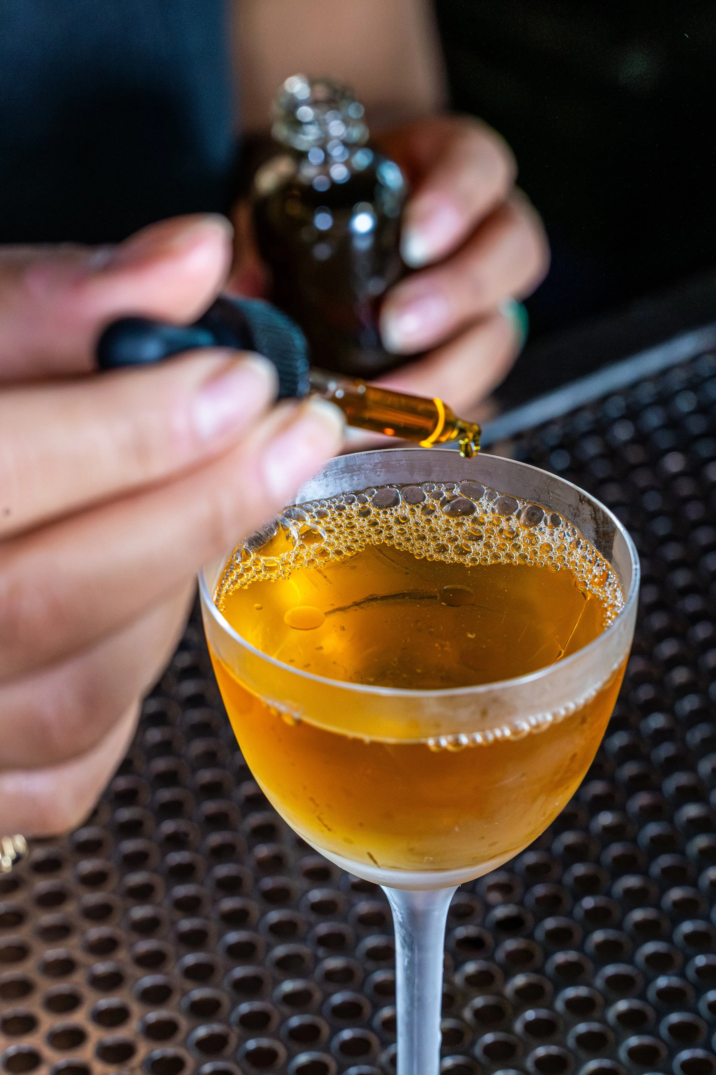 A hand holding a small dark glass bottle with a dropper, with a cocktail glass filled with a golden drink and foam bubbles, placed on a table with a textured surface.