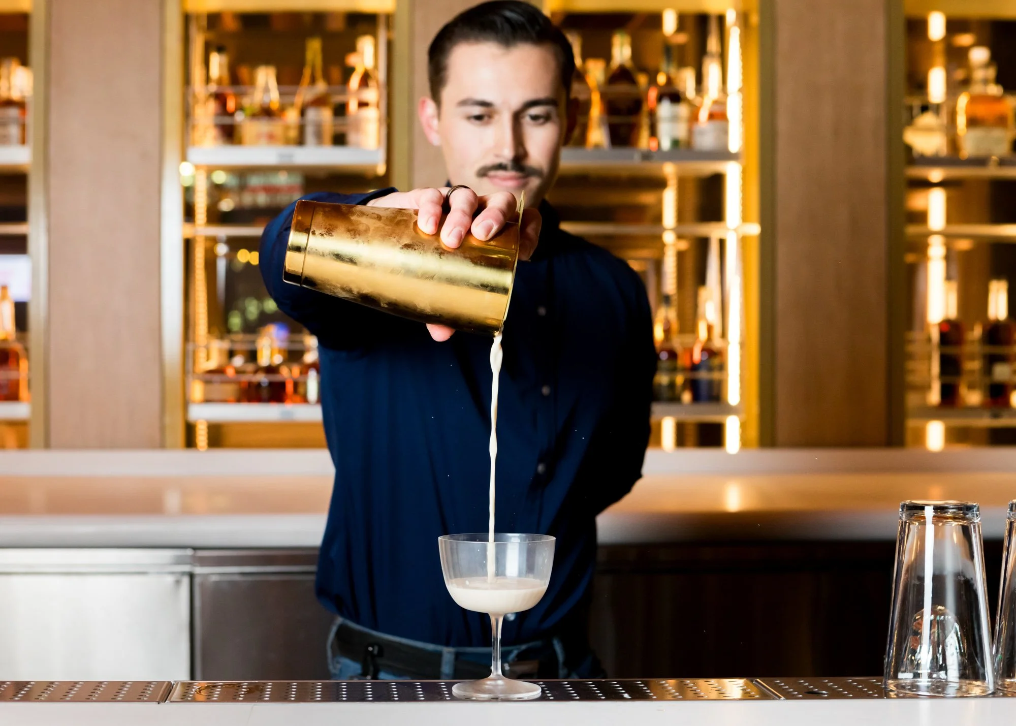 A bartender pours a creamy cocktail from a gold shaker into a coupe glass, with a glass of water and a cocktail shaker on the bar in front of him, in a well-lit bar or restaurant.