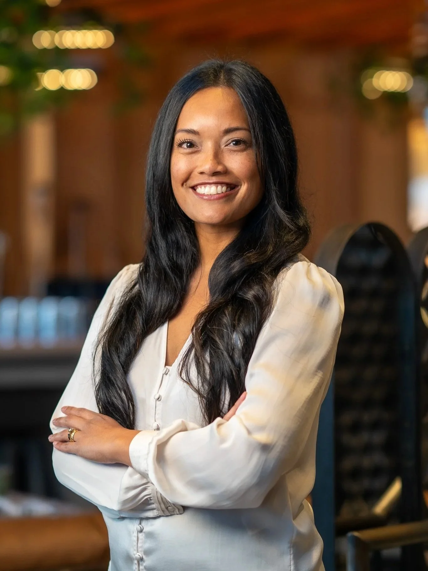 Smiling woman with long dark hair, wearing a cream-colored blouse, standing confidently with arms crossed indoors.