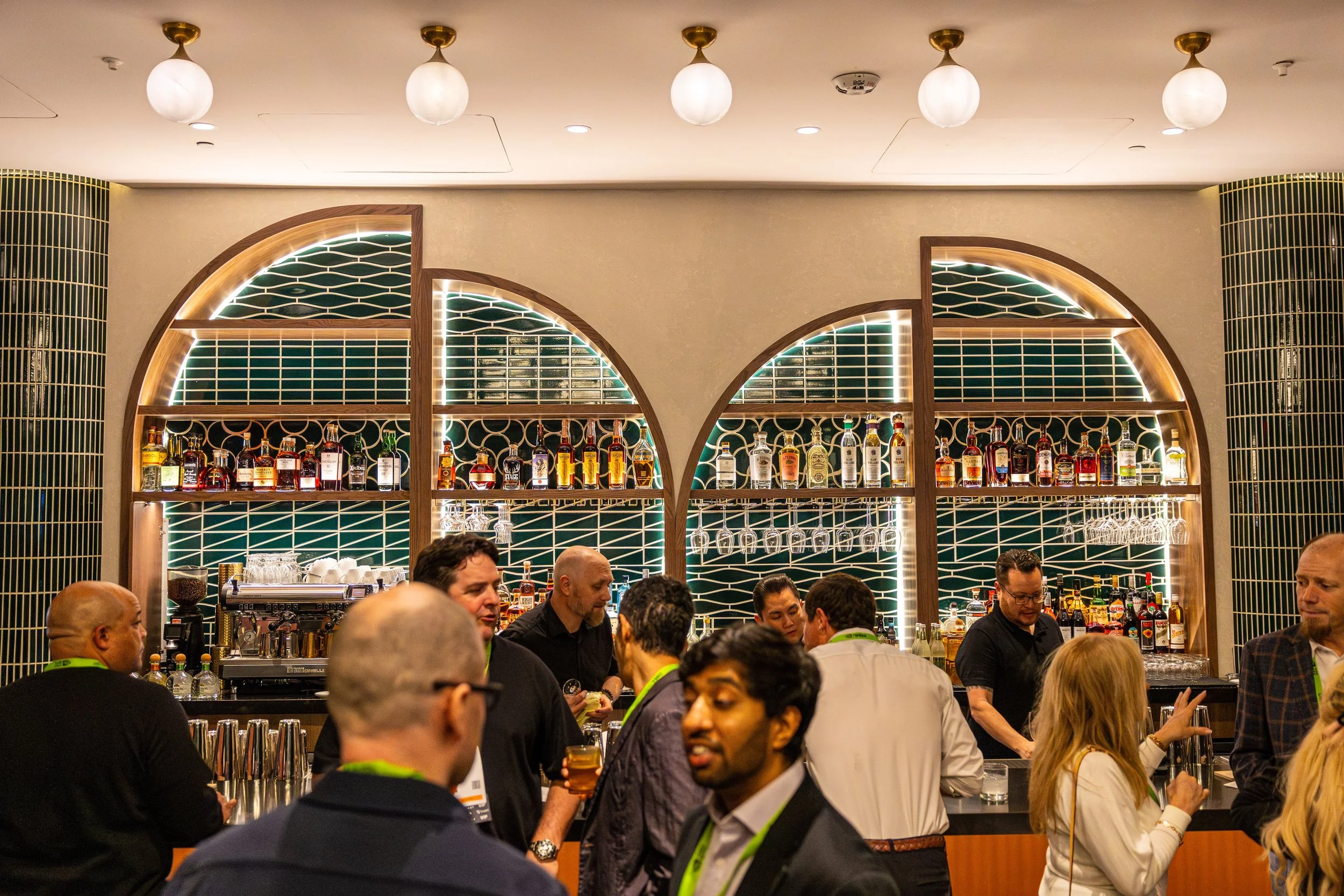 A bar scene with multiple people socializing, behind the bar are shelves with various bottles of alcohol, a coffee machine, and hanging glasses, with decorative green tiles and warm lighting.