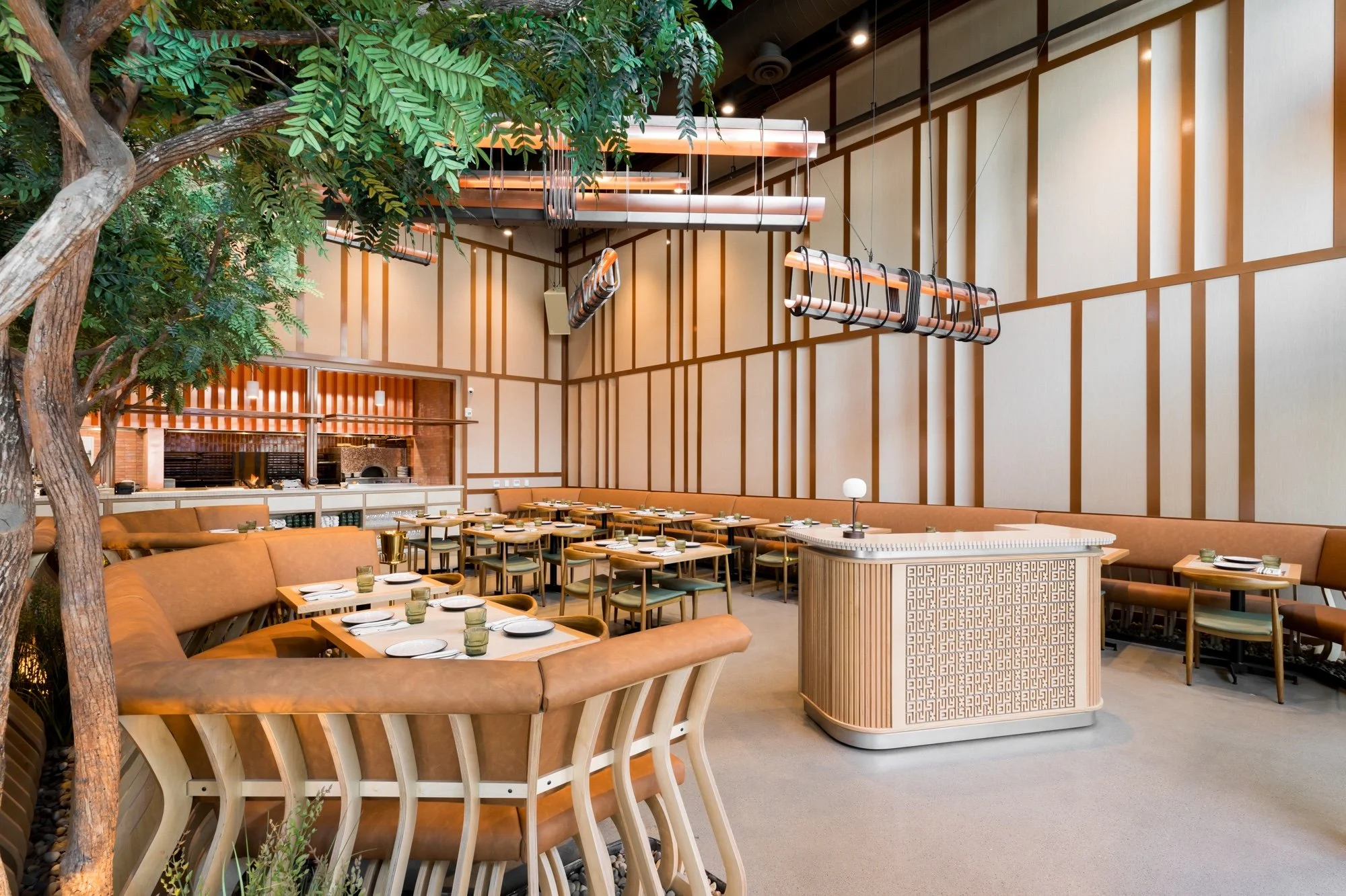 Interior of a modern restaurant with brown and tan decor, tables set with white plates and glasses, a large tree and lush greenery inside, and stylish hanging light fixtures.