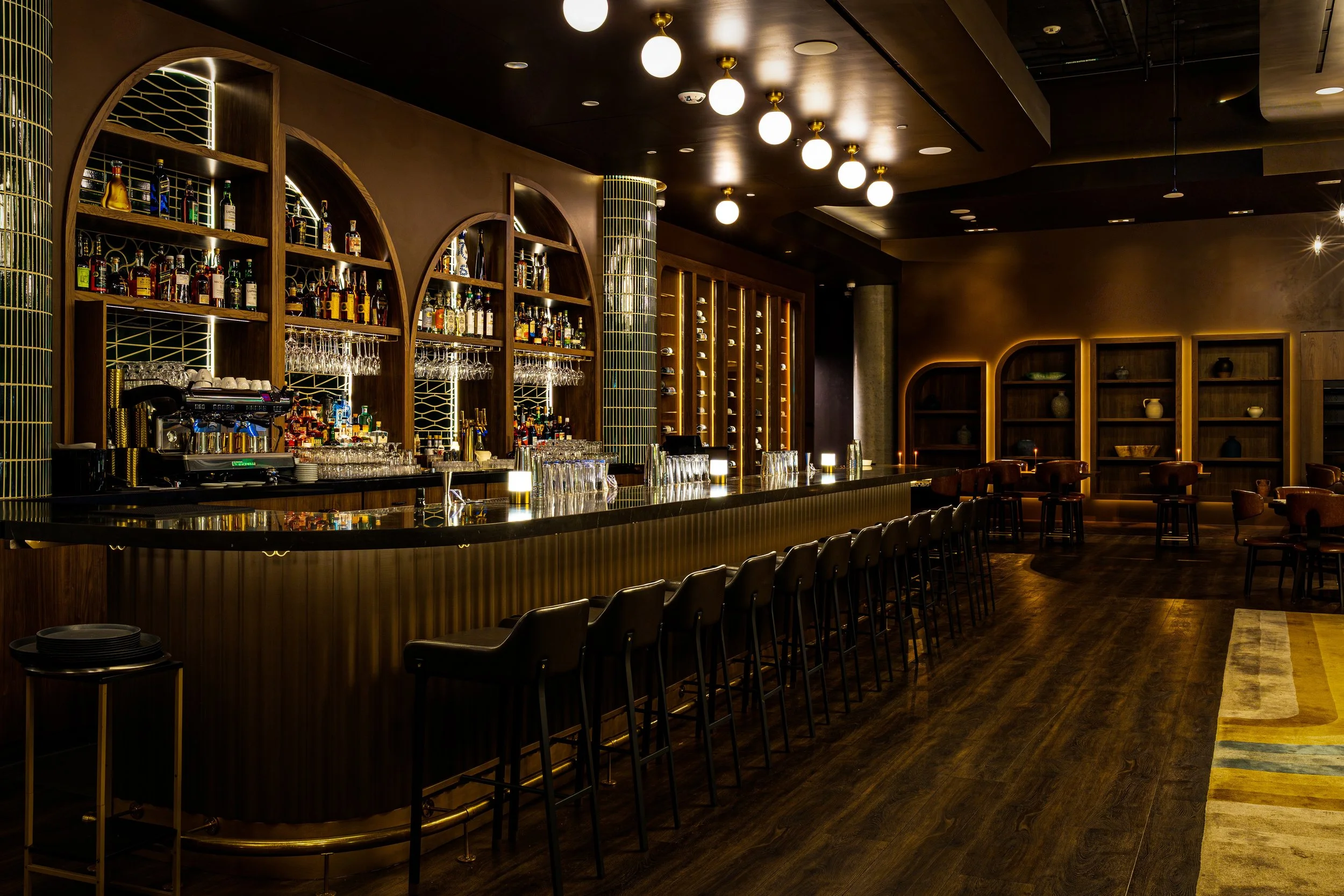 Interior of a modern bar with a long counter, shelves with liquor bottles, glasses, and wine storage, dim lighting, and warm wood accents.