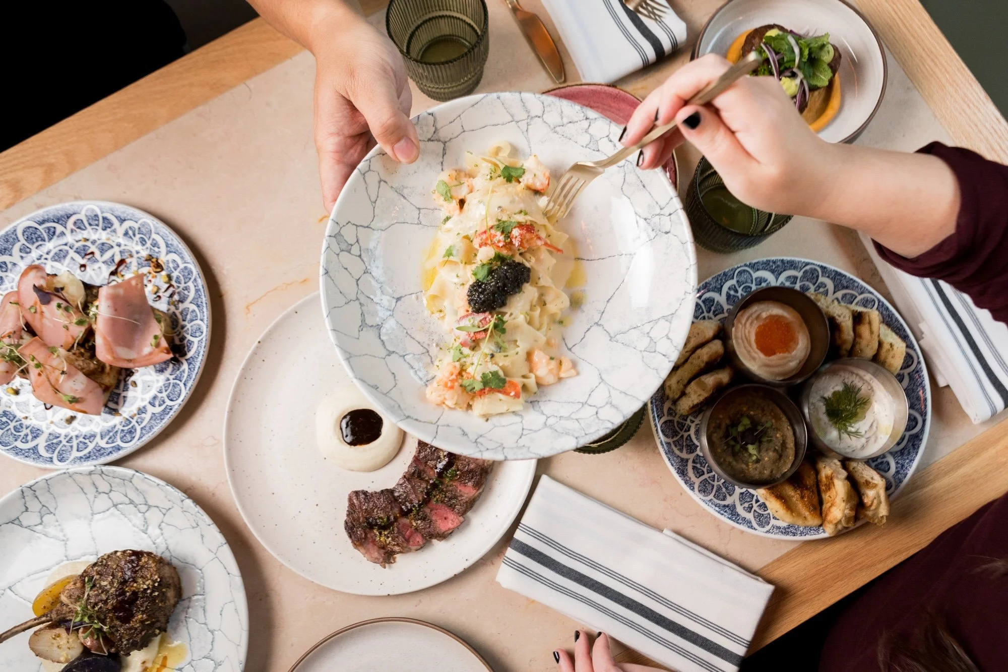 A top-down view of a dinner table with various dishes, including a plate of pasta with seafood, a bowl of salad, grilled meat, and small bowls of dipping sauces, with two hands reaching for the pasta.