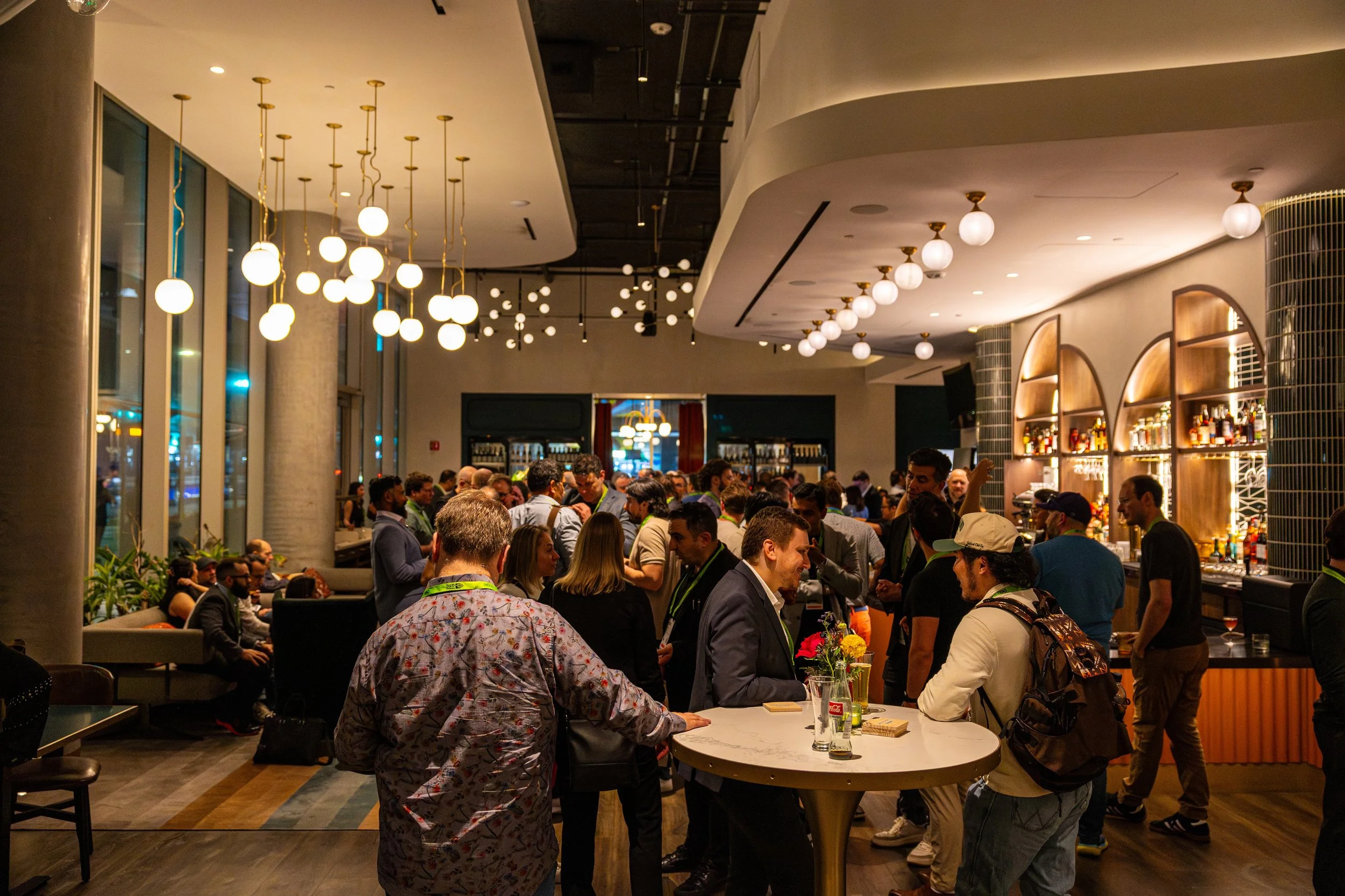Crowd of people socializing and ordering drinks inside a modern bar or restaurant with stylish lighting fixtures and wooden accents.