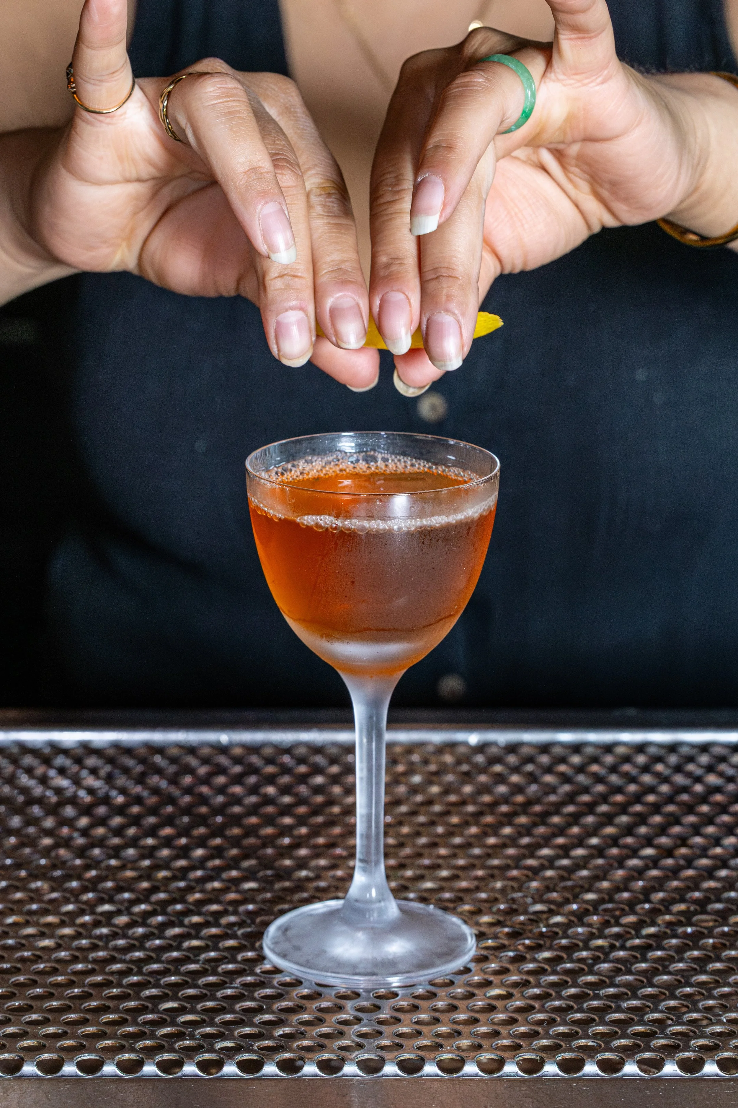 Hands with rings on fingers are squeezing a lemon over a cocktail glass filled with an orange-colored drink.