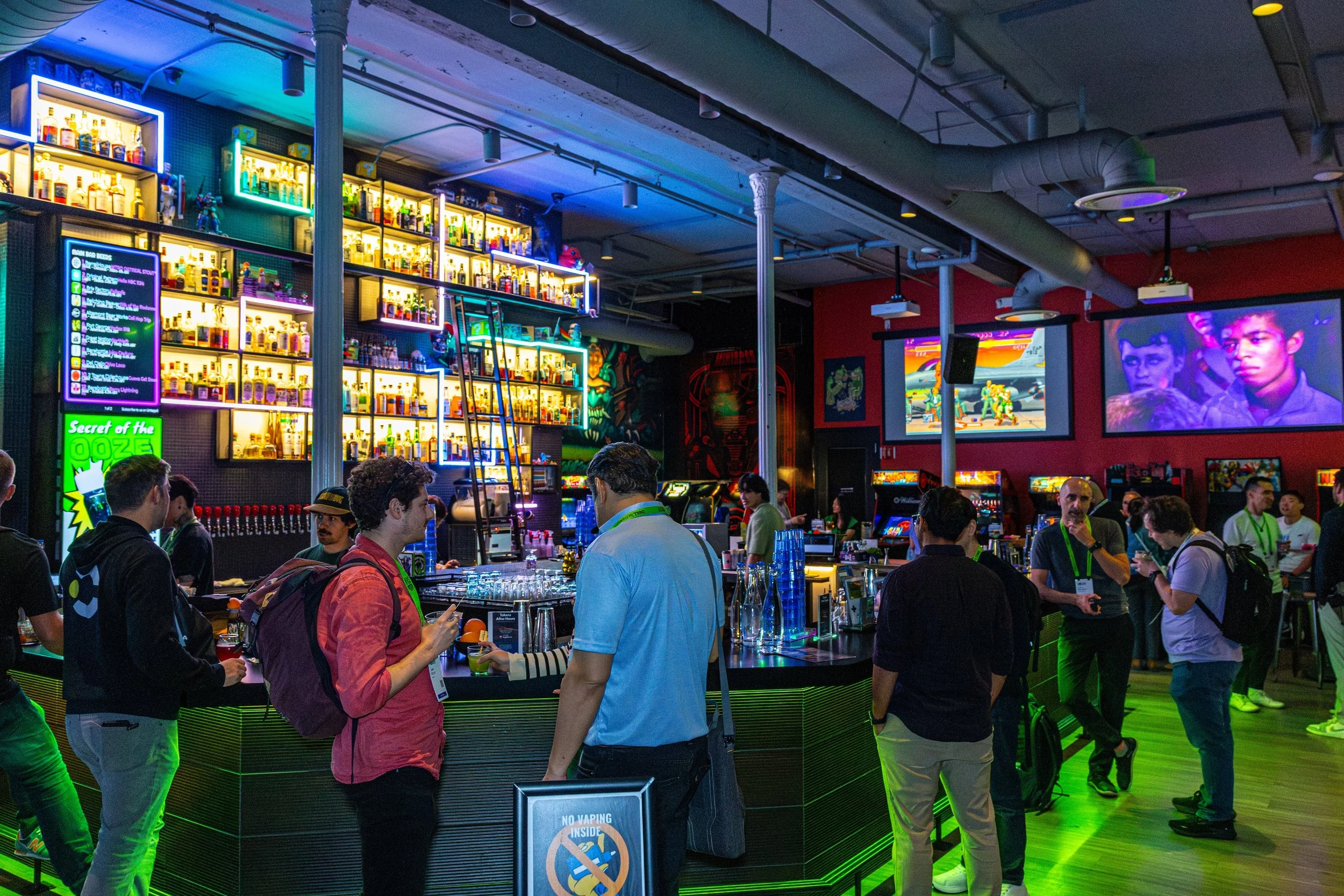 People standing around a bar counter at an indoor event with colorful lighting, shelves of bottles, and screens showing video game characters in the background.