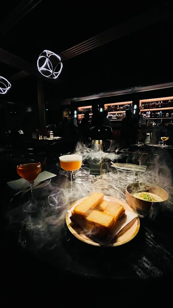 Bar with cocktails and smoked food on the table, illuminated by modern light fixtures, with a dark ambiance and people in the background.