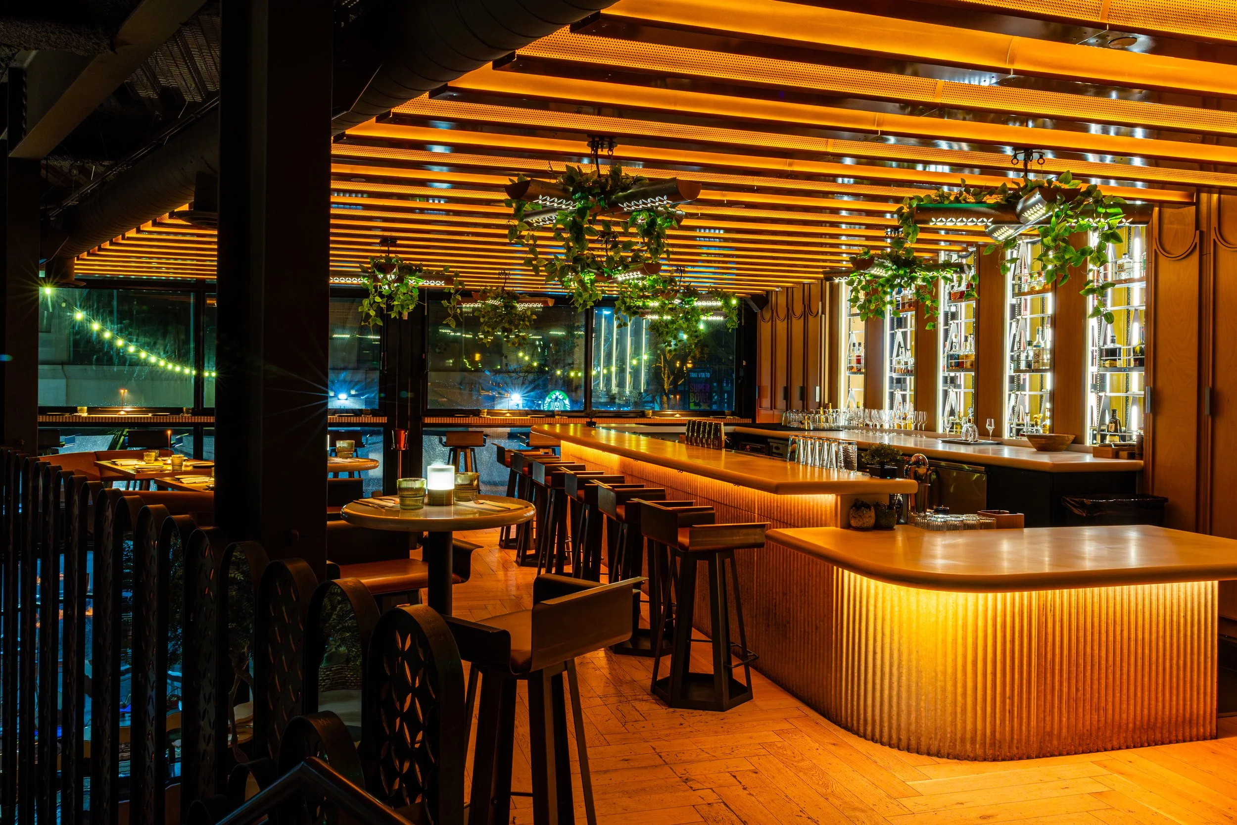Interior of a modern bar or restaurant at night with warm lighting, wooden accents, hanging plants, bar counter with bottles, and city lights visible through large windows.