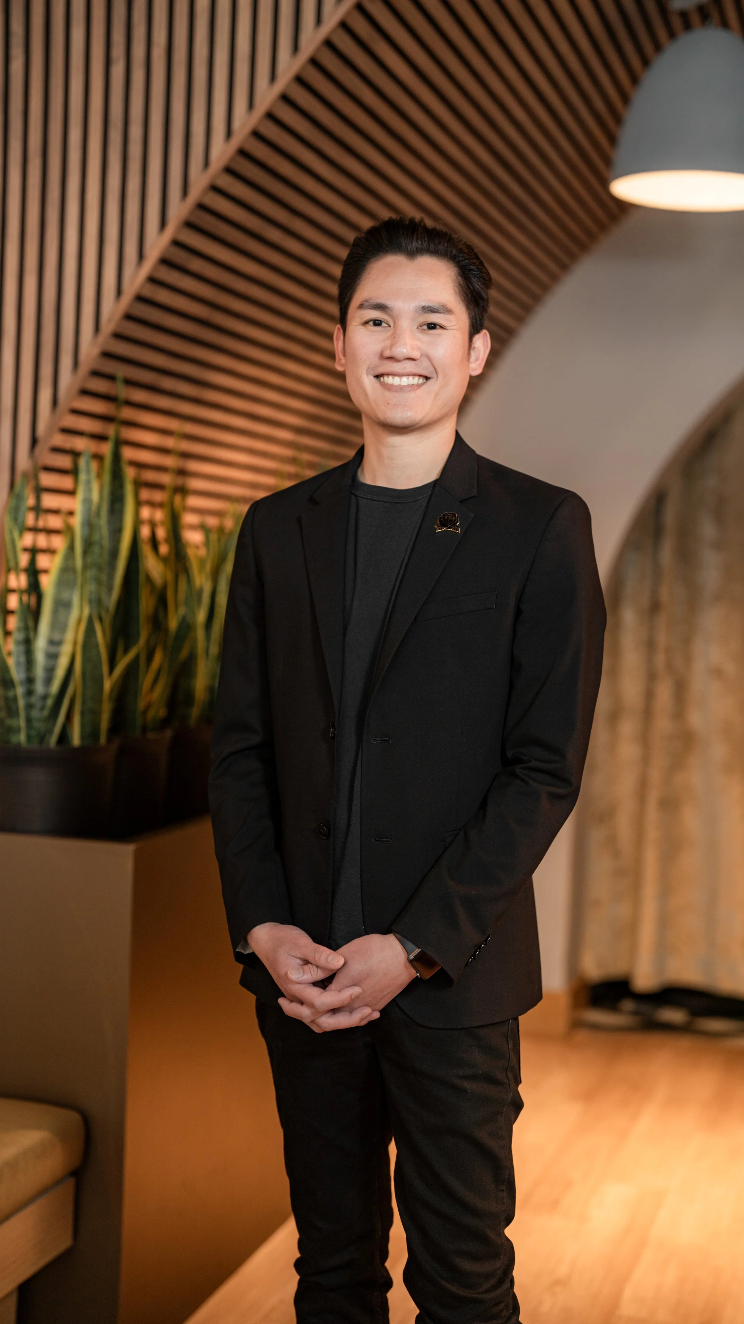 A smiling young man in a black blazer and black pants standing indoors with a wooden slat wall and plants in the background.