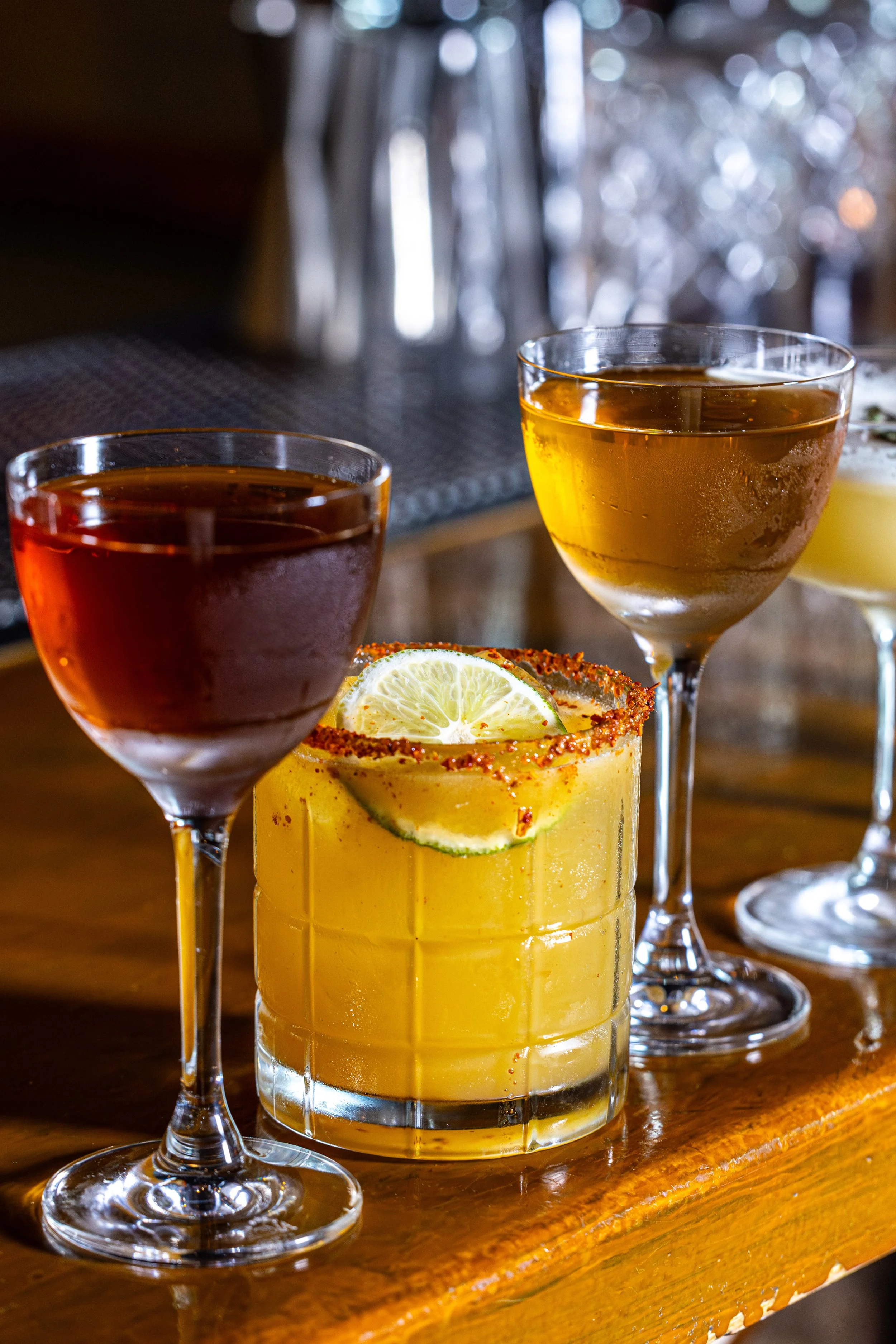 A yellow cocktail drink with lime slices in a tall glass, garnished with a chili powder rim, accompanied by two glasses of red and amber-colored drinks on a wooden surface.