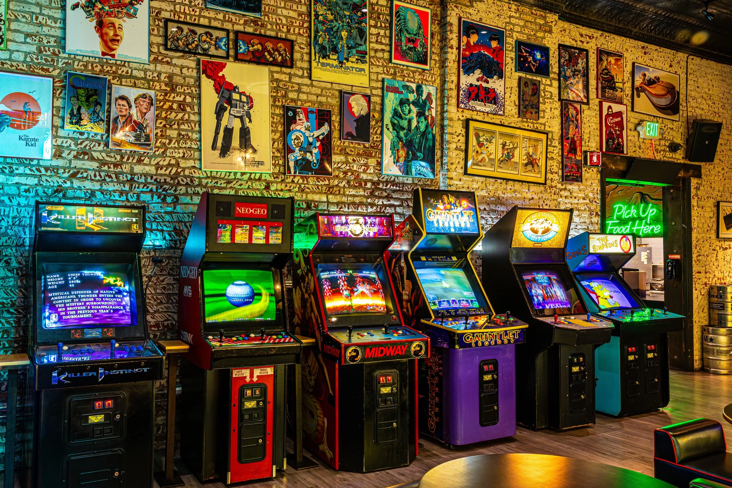 Arcade with multiple vintage video game machines lined up against a brick wall decorated with framed posters of movies, cartoons, and video games. The arcade has neon signs, including a green 'Pick Up Food Here' sign and a 'NEO-GEO' sign. A small rou