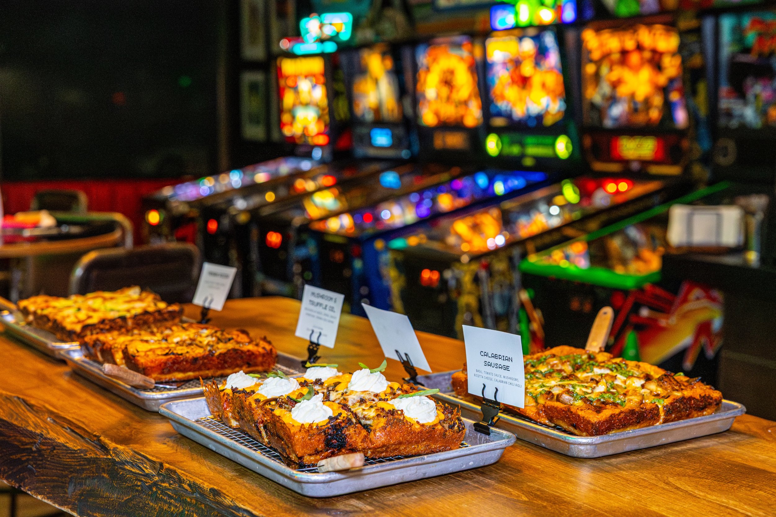 Display of Italian-style baked dishes, including calzone and sausage baking tray, on a wooden table in front of a row of colorful pinball machines in a game room or arcade.