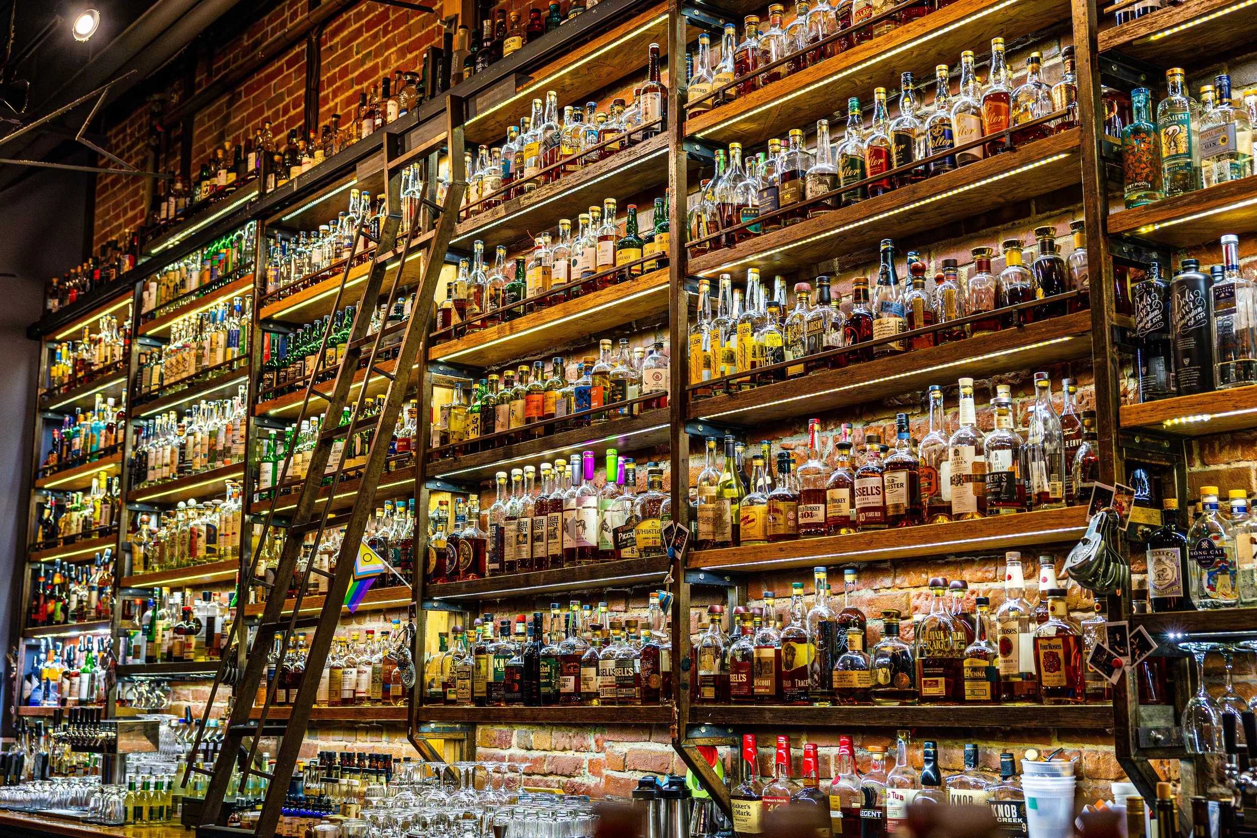 A large wall filled with shelves of various liquor bottles in a bar with brick walls and a ladder to reach higher shelves.