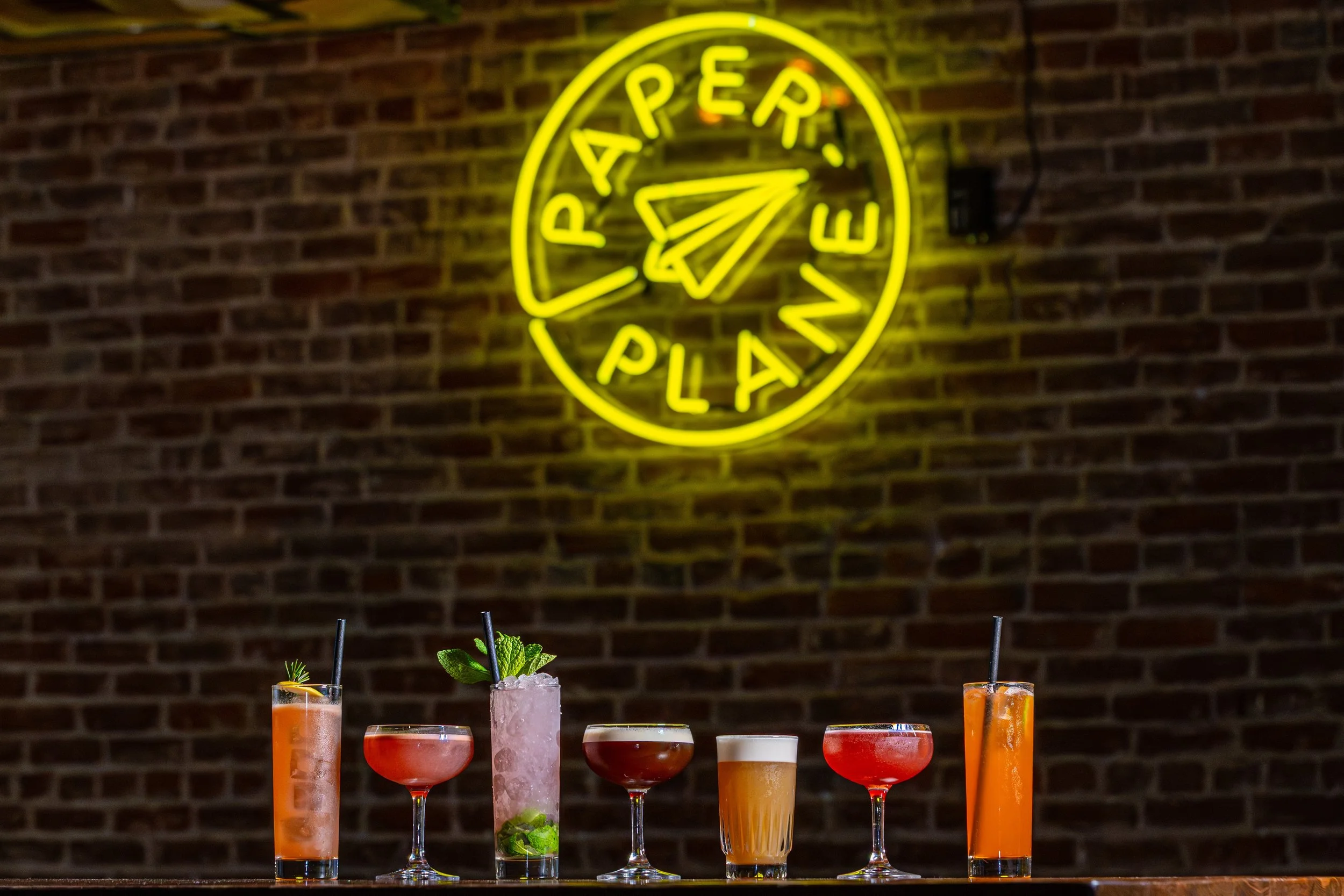 A neon yellow sign on a brick wall reads 'PARTEA PLANE' with a paper plane icon. Below, there are several colorful cocktails in different glasses, garnished with fruits and herbs.