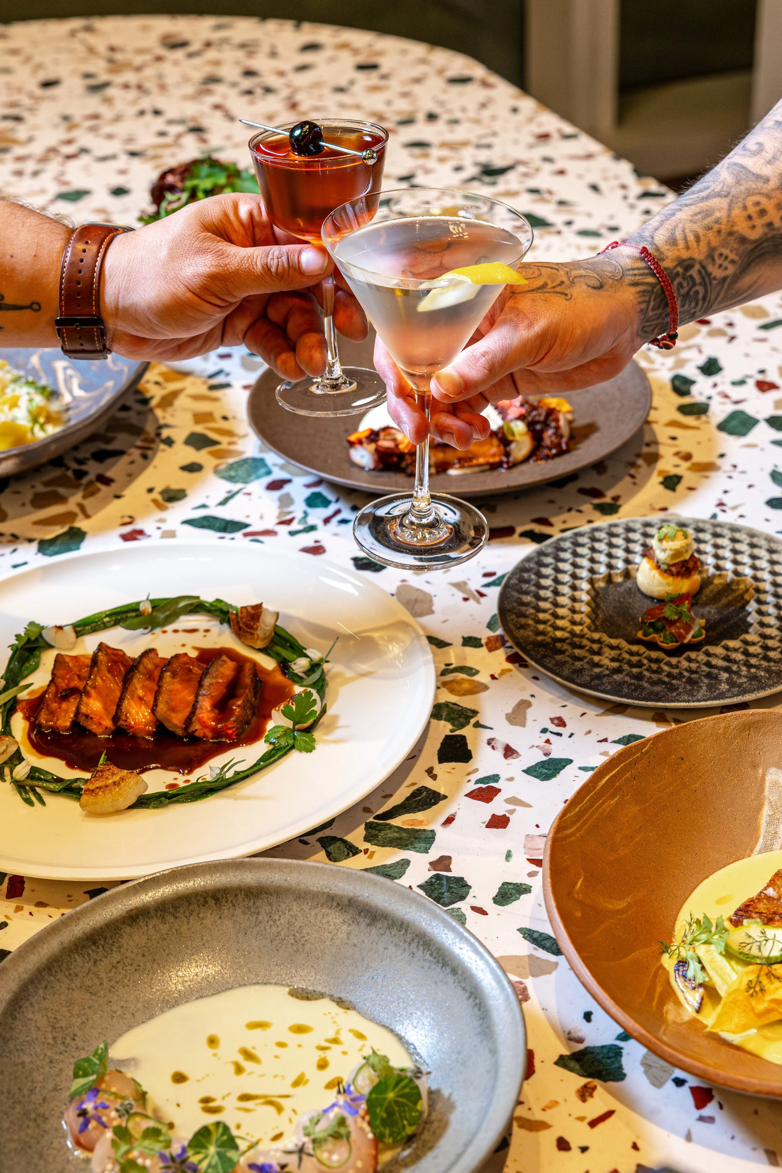 A dinner table with various dishes including sliced meat with sauce, a yellow soup garnished with herbs and edible flowers, and small appetizers with herbs, set on a colorful, patterned tablecloth. Two people are holding cocktail glasses, one with a 