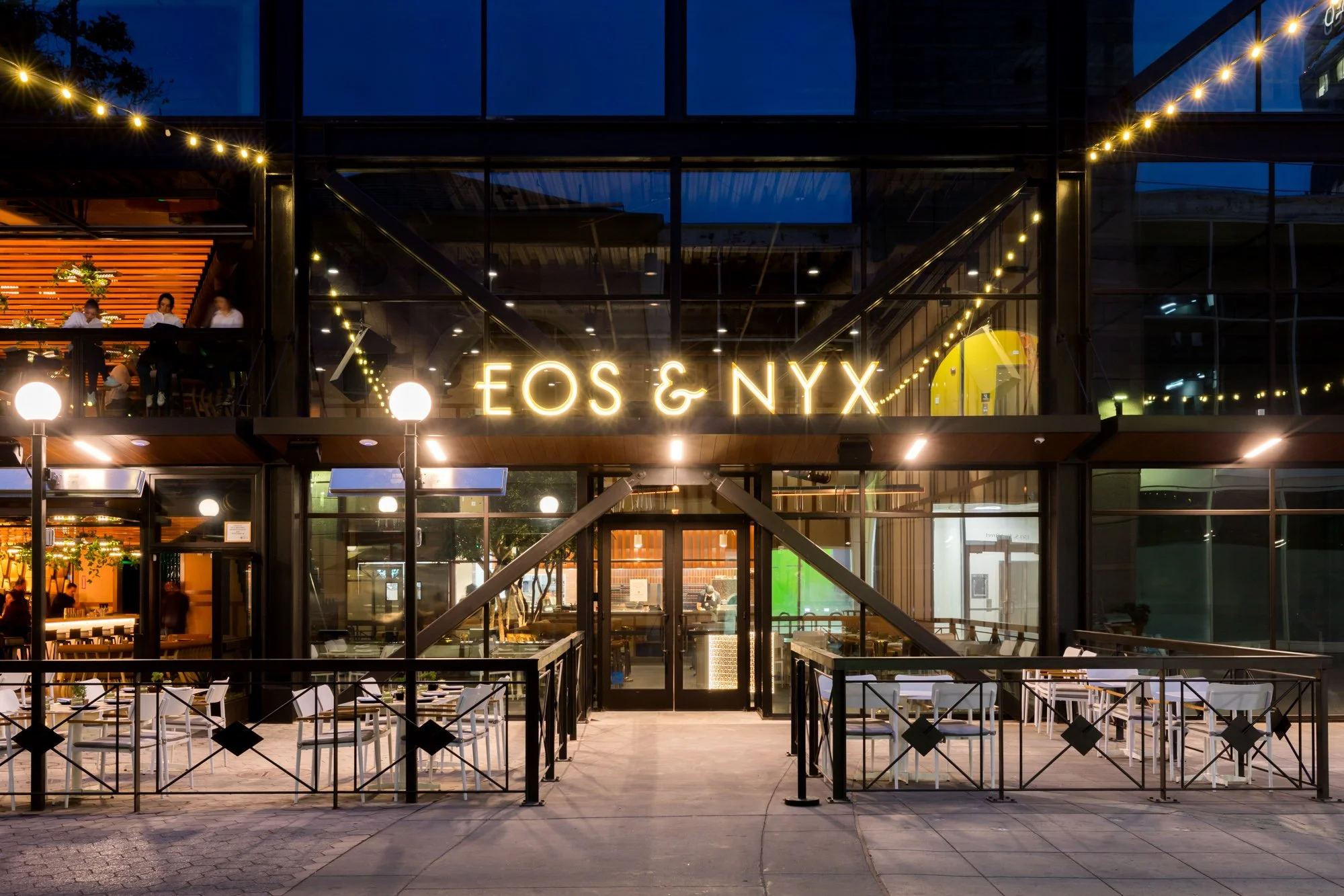 Exterior view of a modern restaurant named EOS & NYX, illuminated at night, with glass walls, outdoor seating, string lights, and a balcony where people are dining.