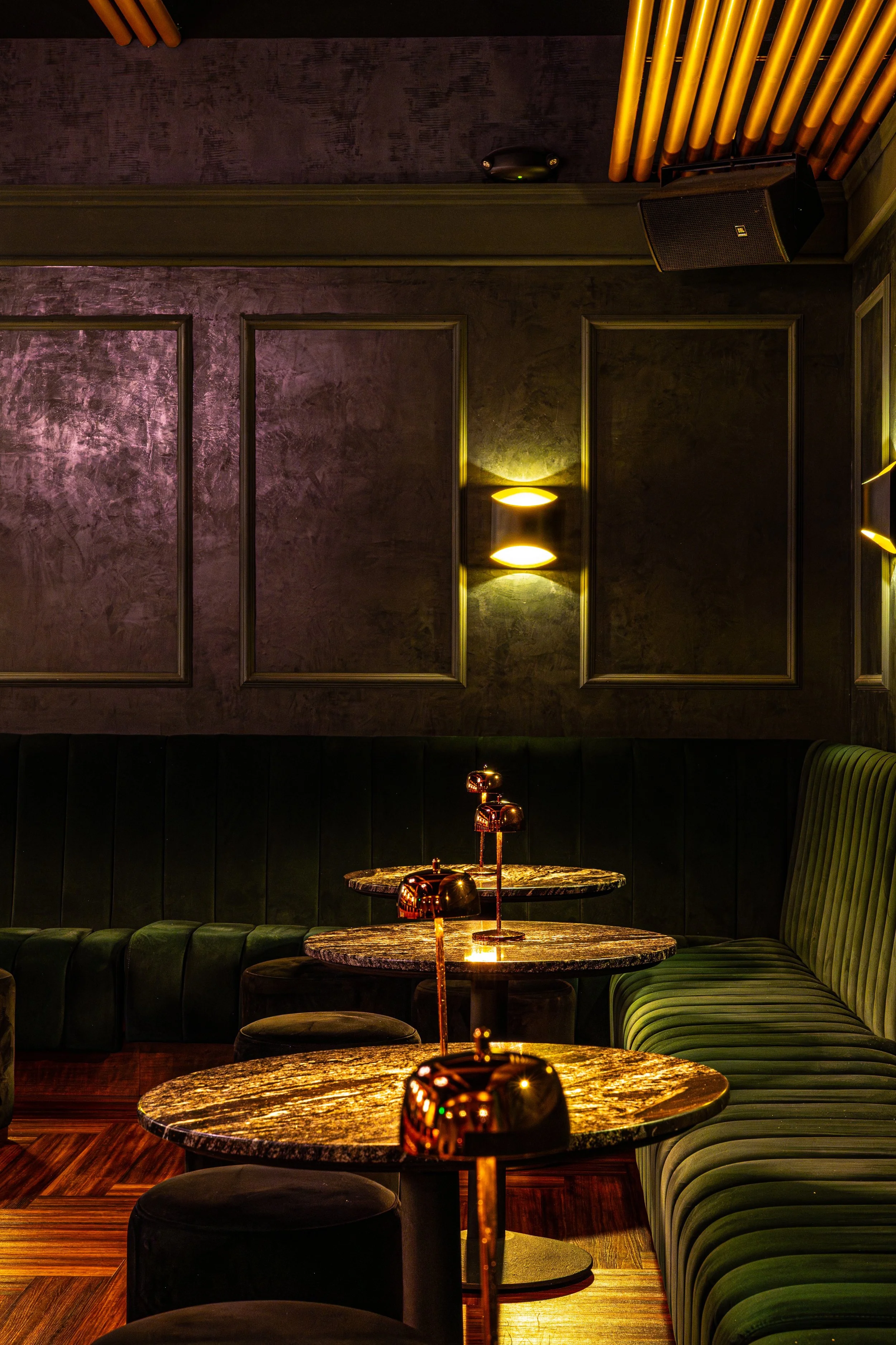 A cozy, dimly-lit restaurant or lounge with rounded marble-top tables and green upholstered banquettes along the wall. Warm yellow lighting enhances the inviting atmosphere.