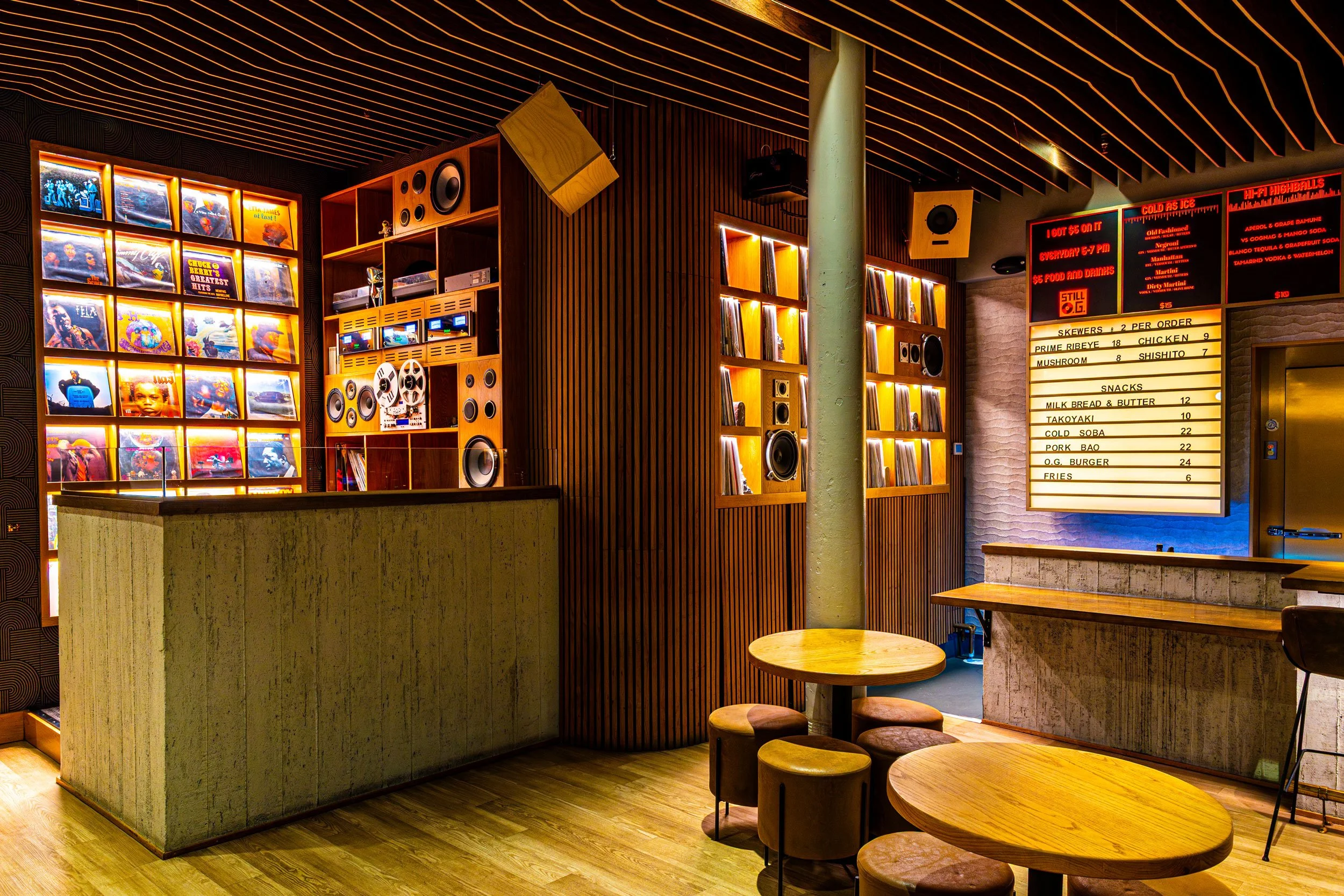 Interior of a modern restaurant or cafe with wooden decor, round tables with matching stools, and a wall featuring illuminated vinyl records and a menu board with drink and food options.