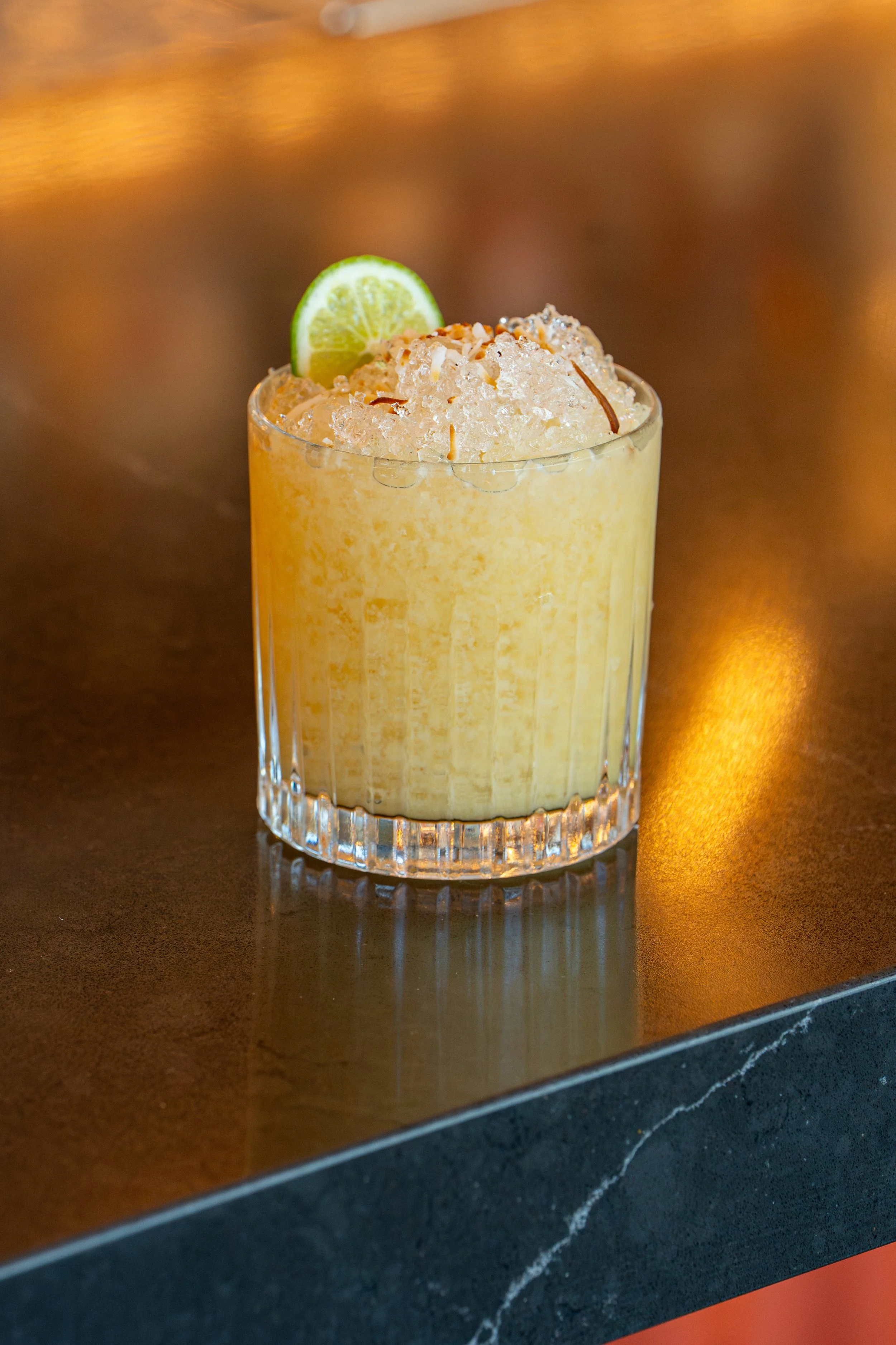 A glass of a yellow frozen drink garnished with a lime wedge, placed on a dark table with a reflective surface.