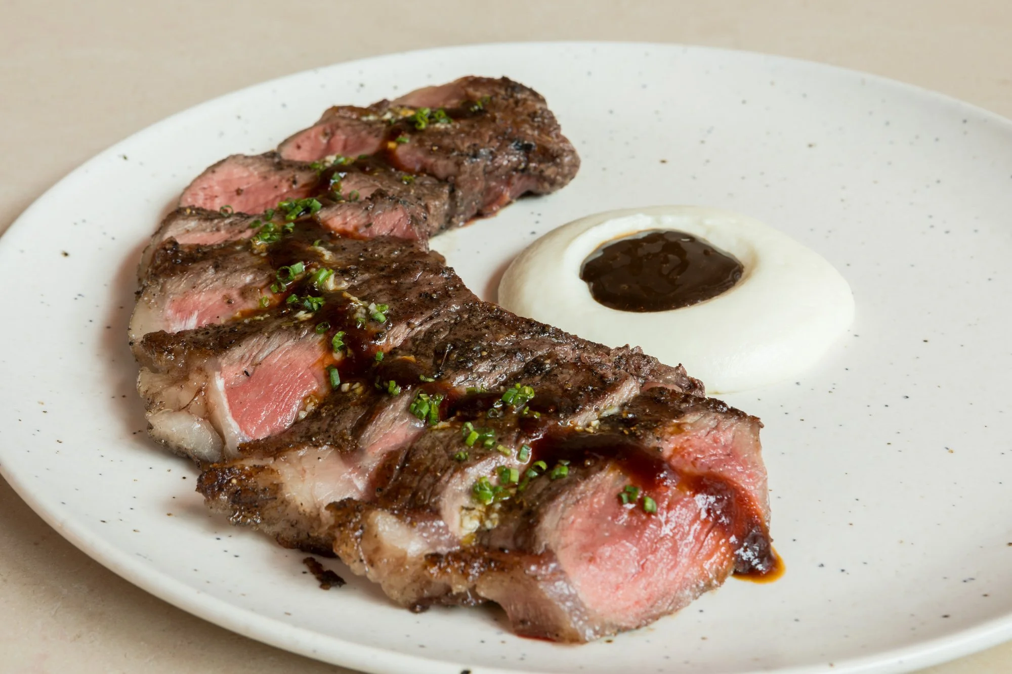 Sliced medium-rare steak topped with chopped chives and sauce, served with a dollop of creamy mashed potatoes with a dark sauce in the center, on a white speckled plate.