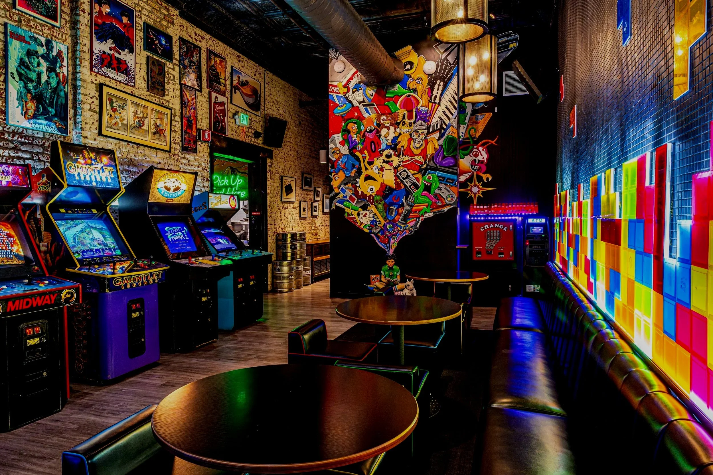 An arcade room decorated with comic and video game-themed artwork on the walls, featuring vintage arcade machines, colorful graffiti, and neon signs. There are tables and chairs for seating.
